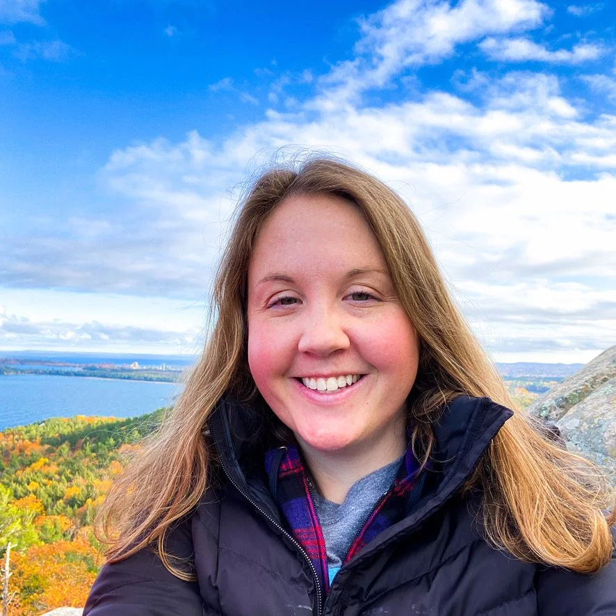 A woman smiling outdoors with fall foliage, a large body of water, and a cloudy sky in the background.