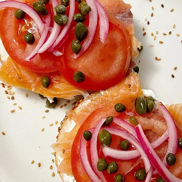 Close-up of smoked salmon canapés topped with tomato slices, sliced red onions, green capers, and sprinkled sesame seeds on a white plate.
