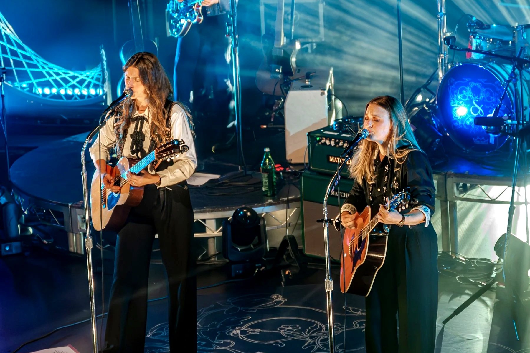Two women performing live on stage with guitars. The woman on the left has long wavy hair, is wearing a light-colored blouse with a black ribbon, and is singing into a microphone while playing an acoustic guitar. The woman on the right has long straight hair, is wearing a black outfit, and is also singing into a microphone while playing an acoustic guitar. Stage lighting and musical equipment are visible in the background.