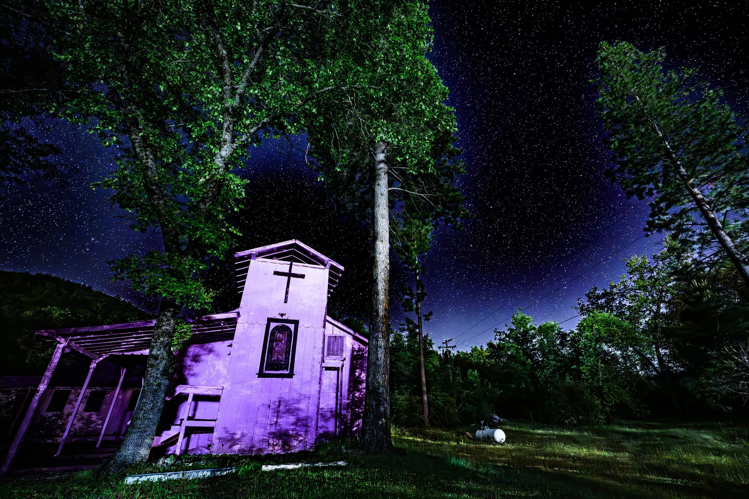 Night sky filled with stars above an old, weathered church illuminated with purple light, surrounded by tall trees and utility poles.