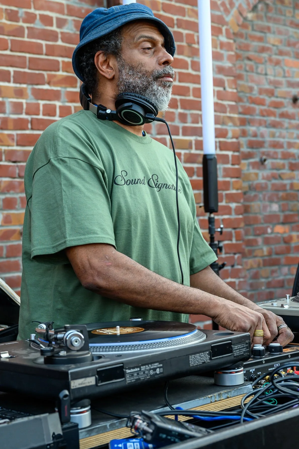 A DJ wearing a blue hat and green t-shirt with 'Sound Signature' logo, standing outdoors against a brick wall, operating DJ equipment with headphones around his neck.