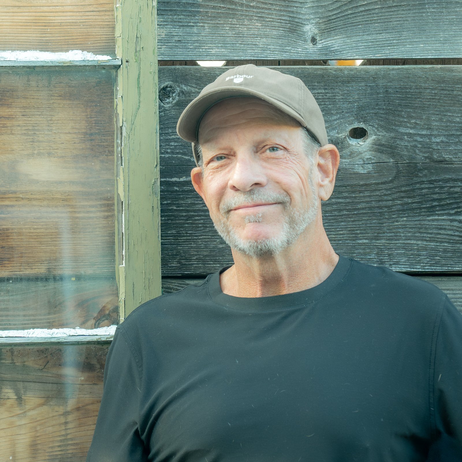 A middle-aged man with light skin, gray hair, and a gray beard wearing a beige baseball cap and a black shirt, standing outdoors against a wooden fence.
