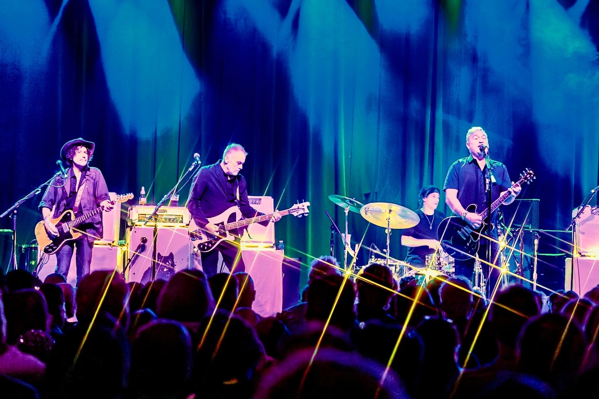 A band performs on stage with colorful lighting and laser effects, with audience visible in the foreground.