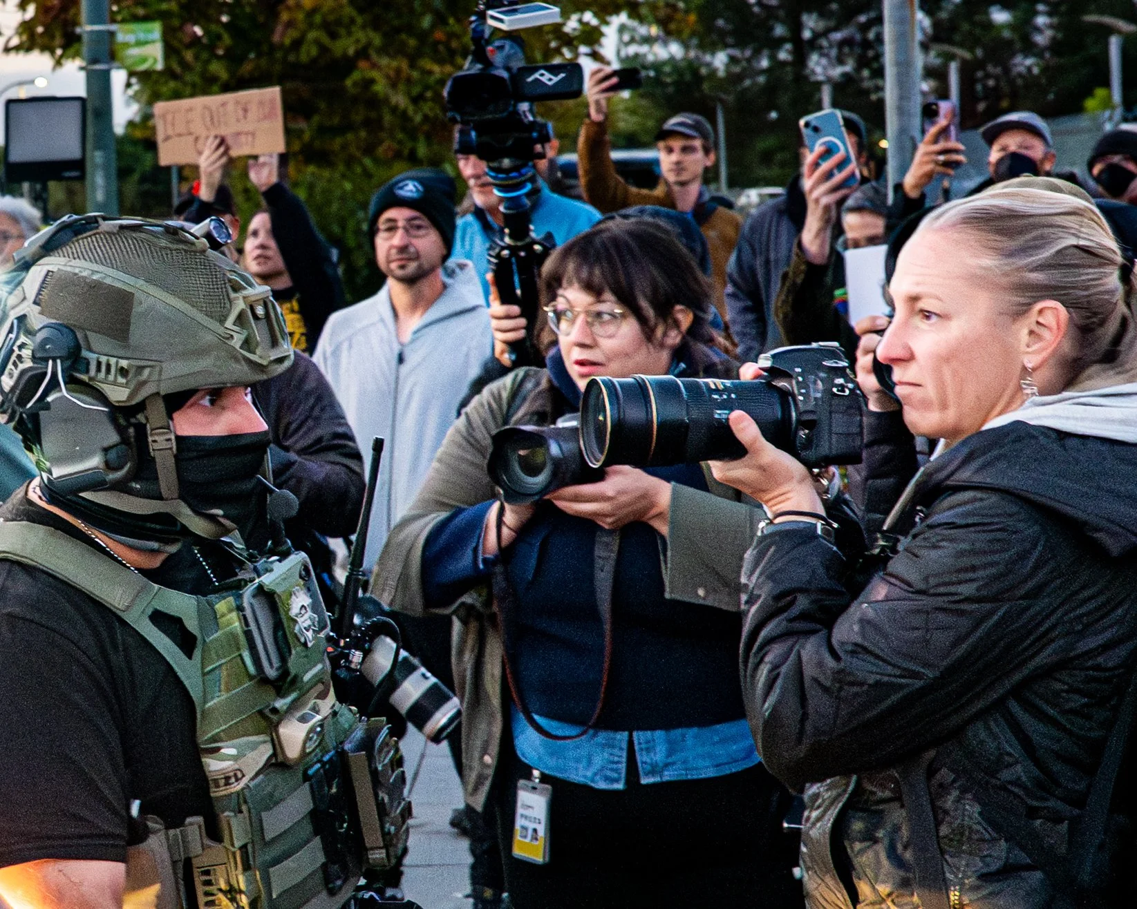 Police officer in tactical gear facing a woman with a camera during a protest, with protesters and cameras in the background.