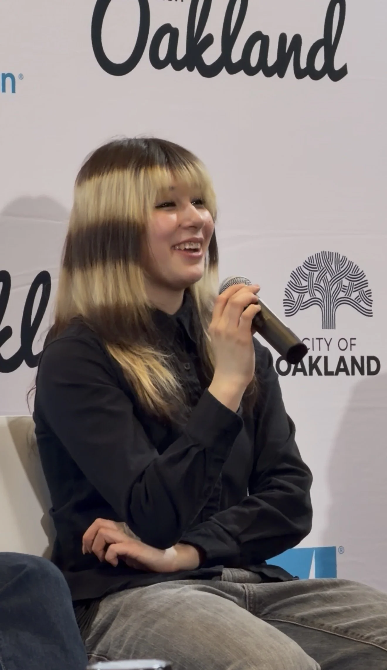A young woman with long blonde hair, wearing a black shirt and gray pants, smiling and holding a microphone at an Oakland event, with a backdrop displaying the words 'Oakland' and a logo that says 'City of Oakland'.
