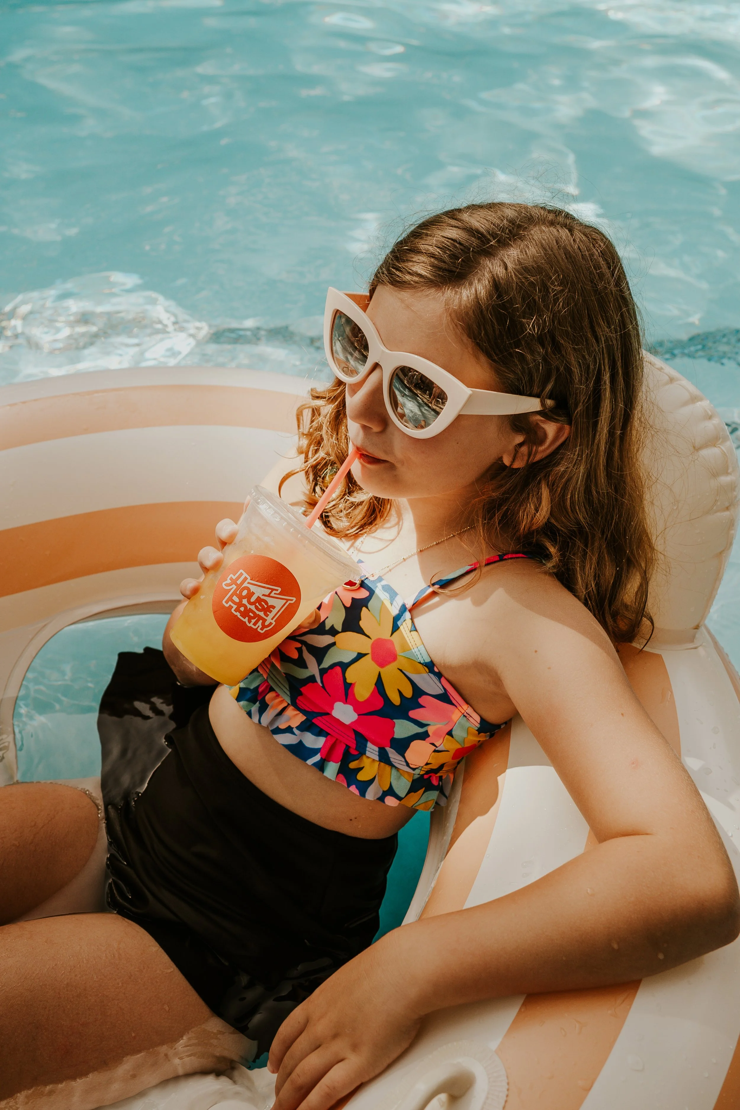 A girl with curly brown hair wearing sunglasses, a colorful floral swimsuit, and black shorts, relaxing on a float in a swimming pool while drinking a beverage through a straw.