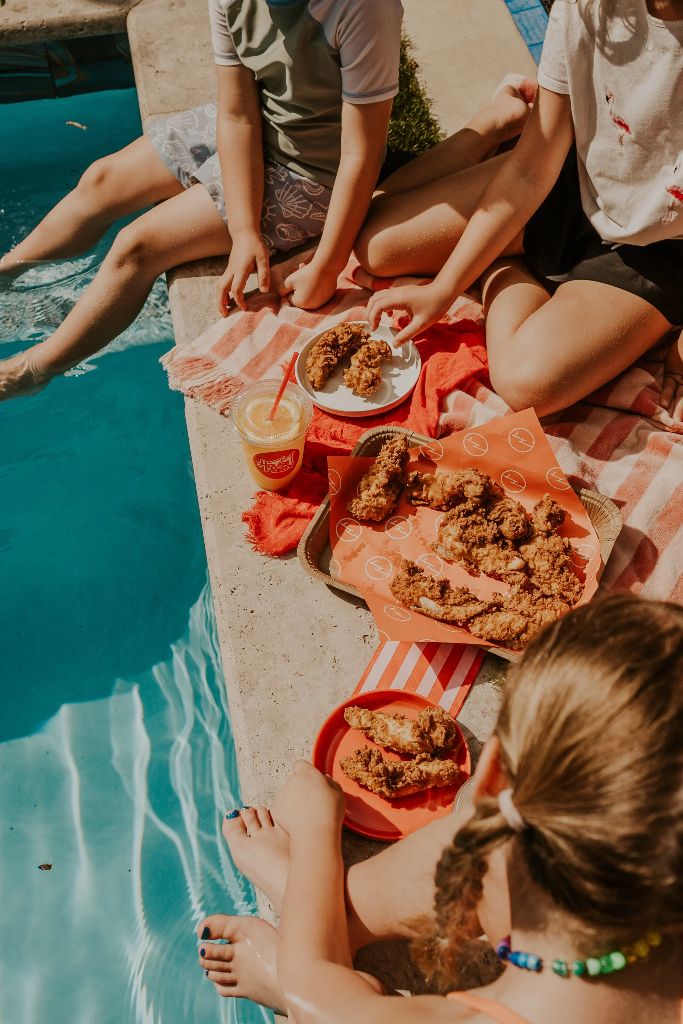 Children sitting by a poolside with fried chicken meal and a drink, with one child dipping their toes in the pool.