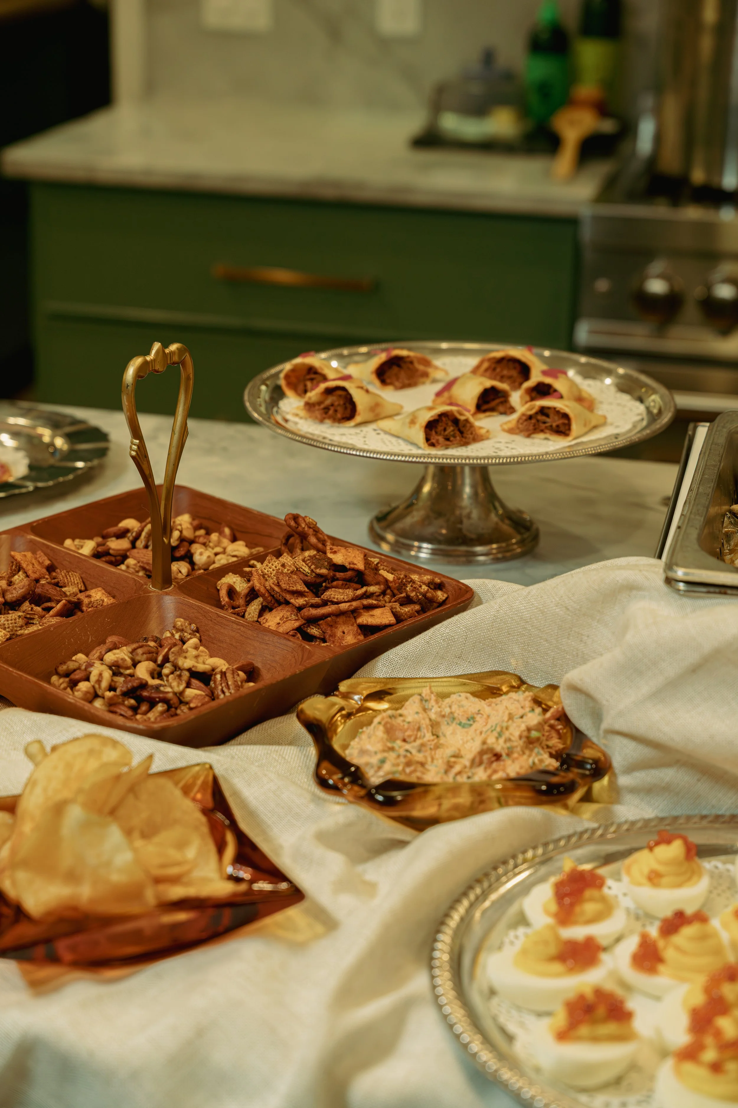 A table with various desserts including stuffed pastry, assorted nuts, dip, and small appetizers on white cloth, with a kitchen in the background.