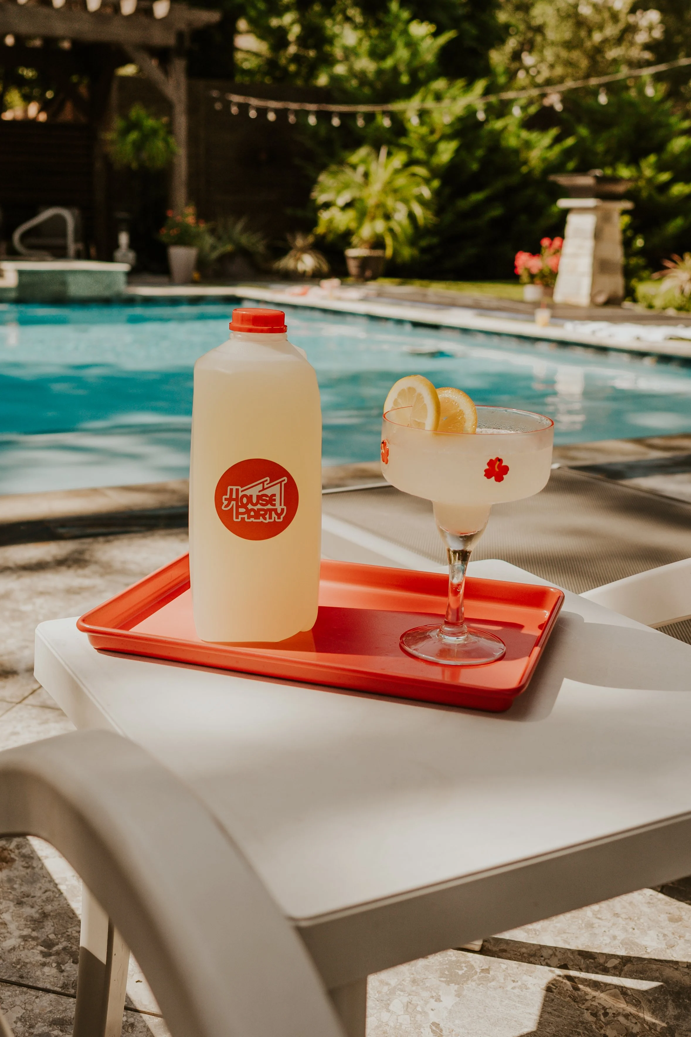 Poolside scene with a tray holding a bottle of sunscreen and a cocktail garnished with lemon slices on a white plastic table in front of a swimming pool, surrounded by greenery and outdoor furniture.