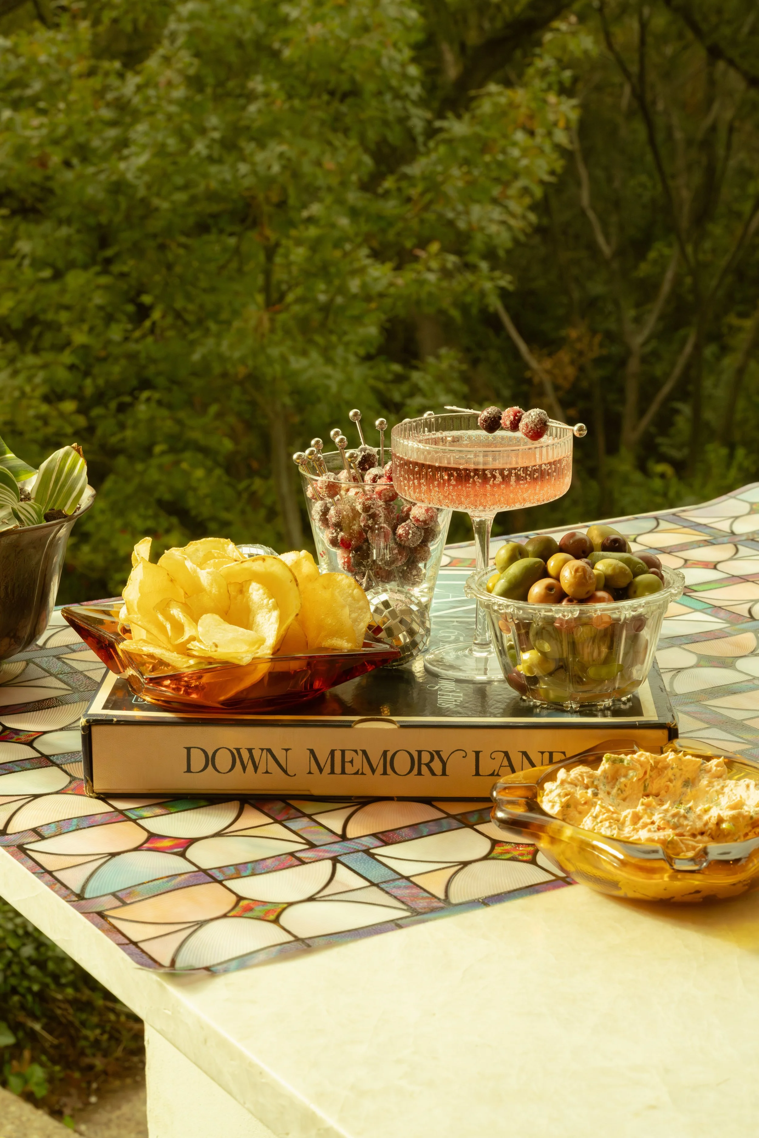 A table set with chips, grapes, a pink cocktail, and dip, with a green forest background.
