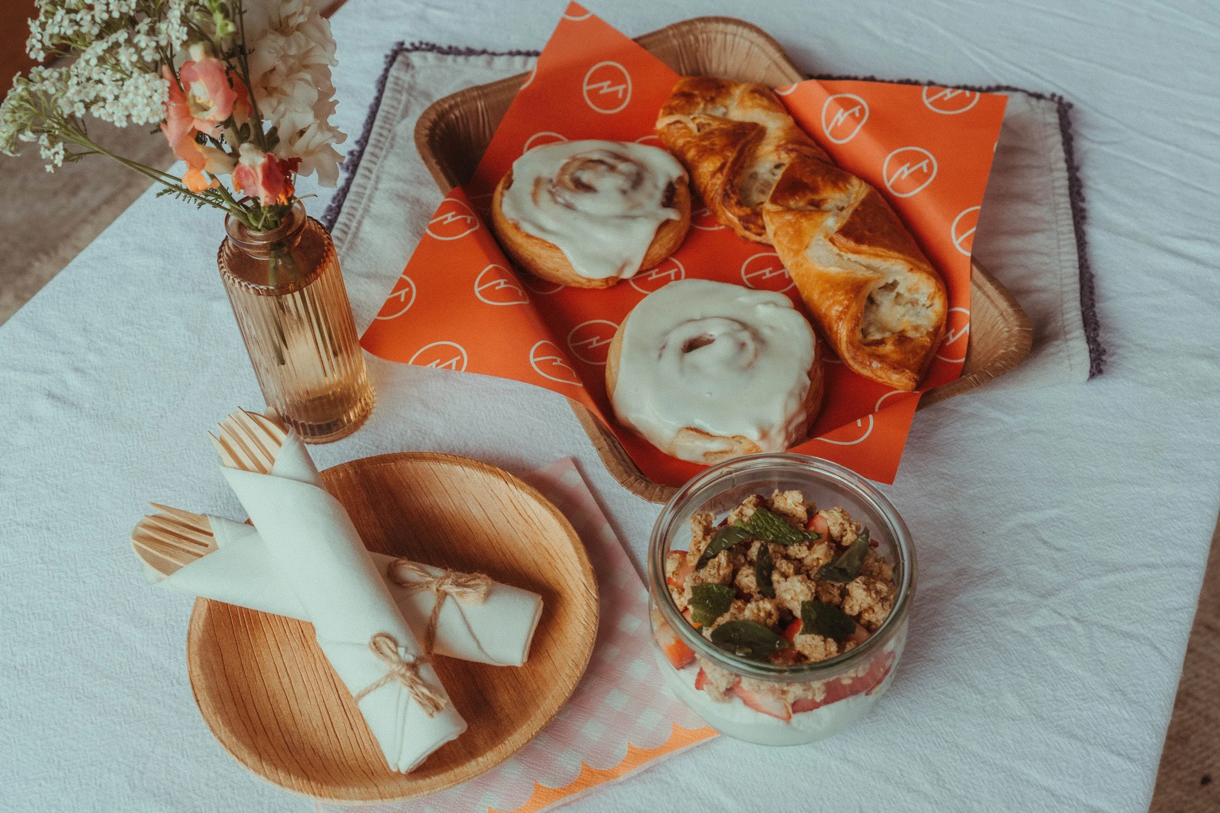 A tray with a cinnamon roll and a croissant, both topped with icing, on an orange patterned paper. To the left, a tall vase with pink and white flowers. In the foreground, there is a small wooden plate with a wrapped napkin.