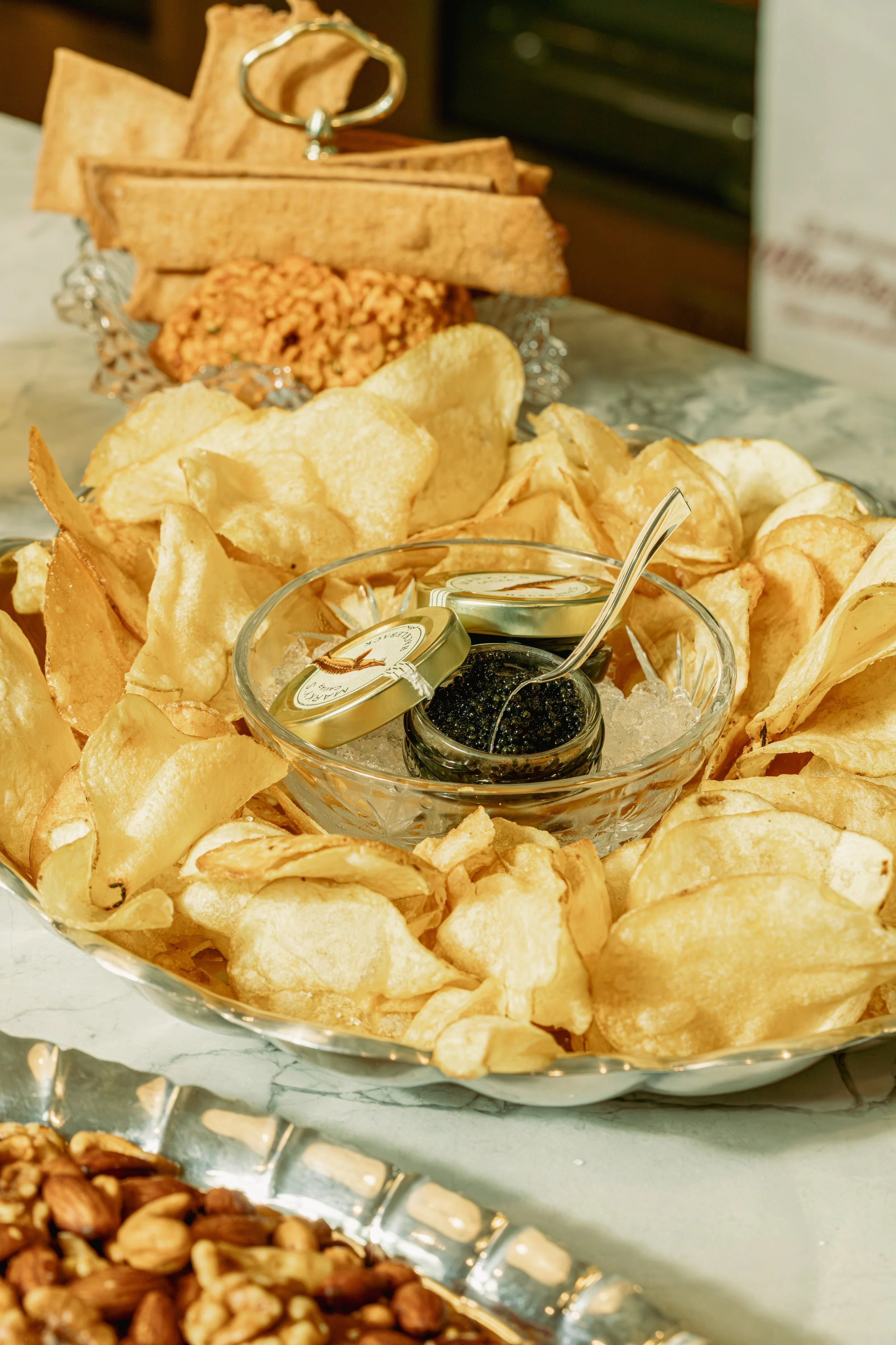 A silver tray filled with potato chips and two small jars of caviar, one with a spoon, surrounded by other snack foods on a marble surface.