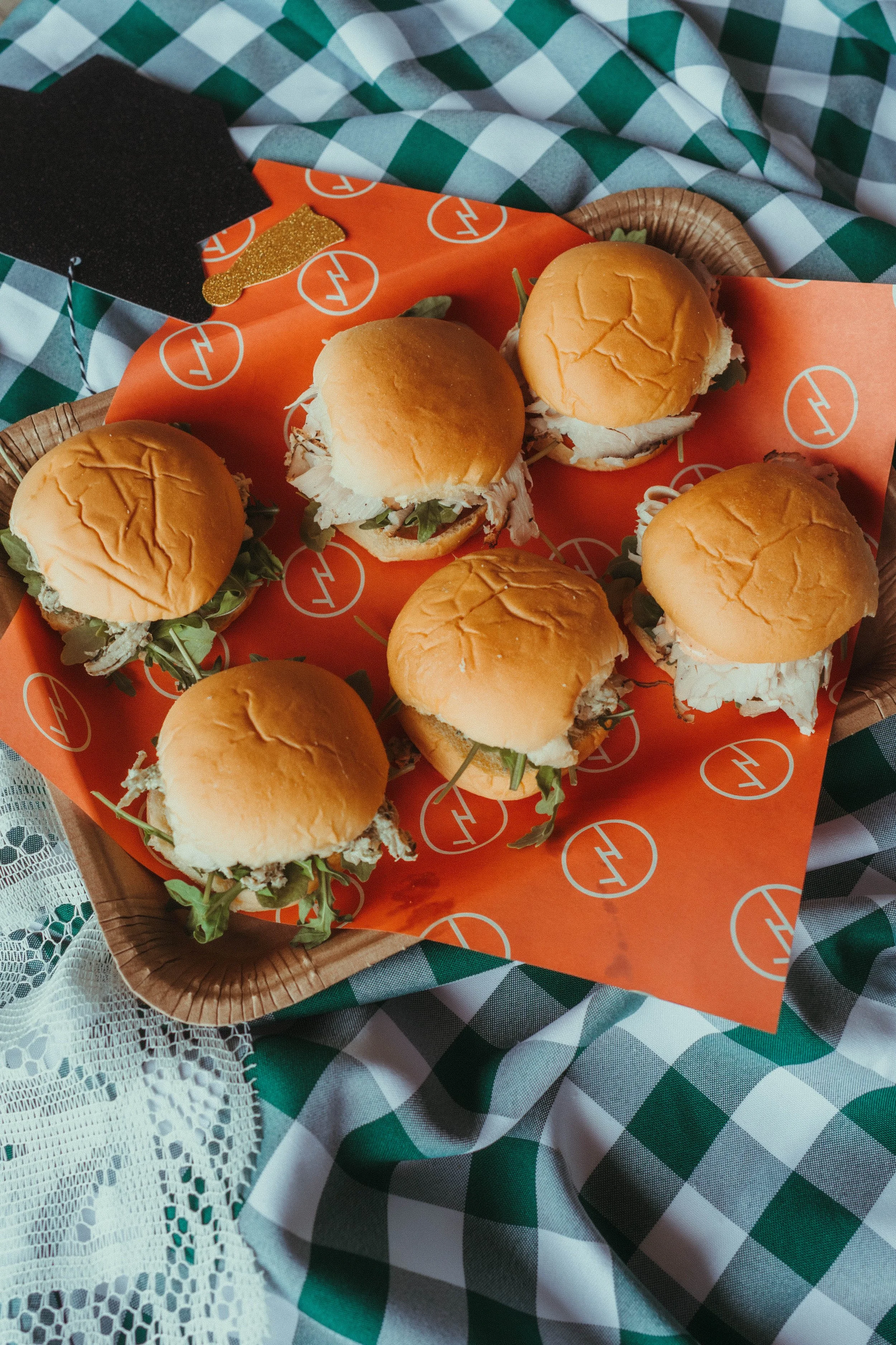 A basket of seven chicken sandwiches with buns, shredded chicken, lettuce, and sauce, placed on orange paper with a white geometric logo at a picnic table with checkered and lace tablecloths.