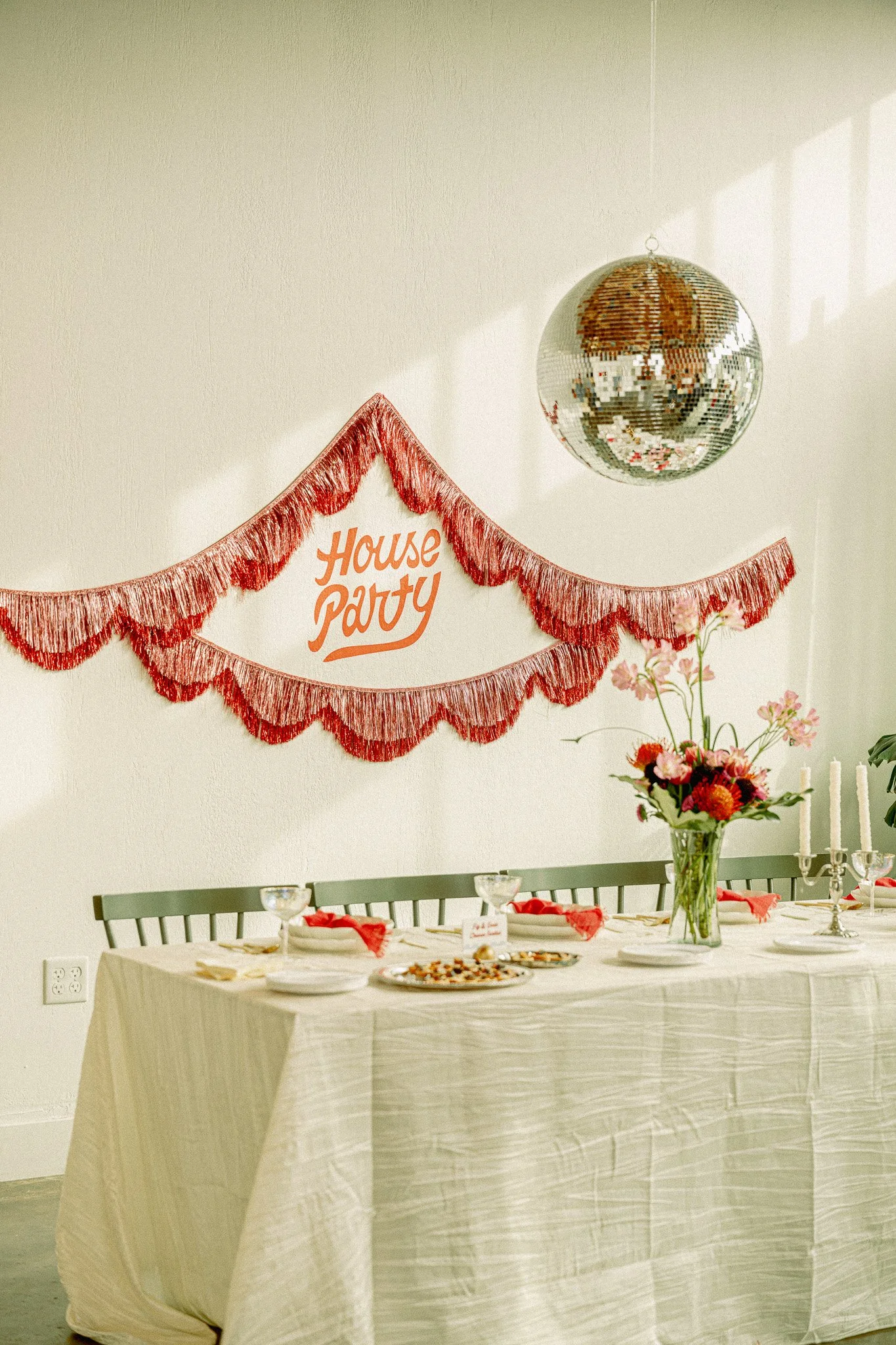 A decorated party space with a banner that reads "House Party," a disco ball hanging from the ceiling, a table set with food and drinks, a vase with pink flowers, and candles, suggesting a celebration or birthday event.
