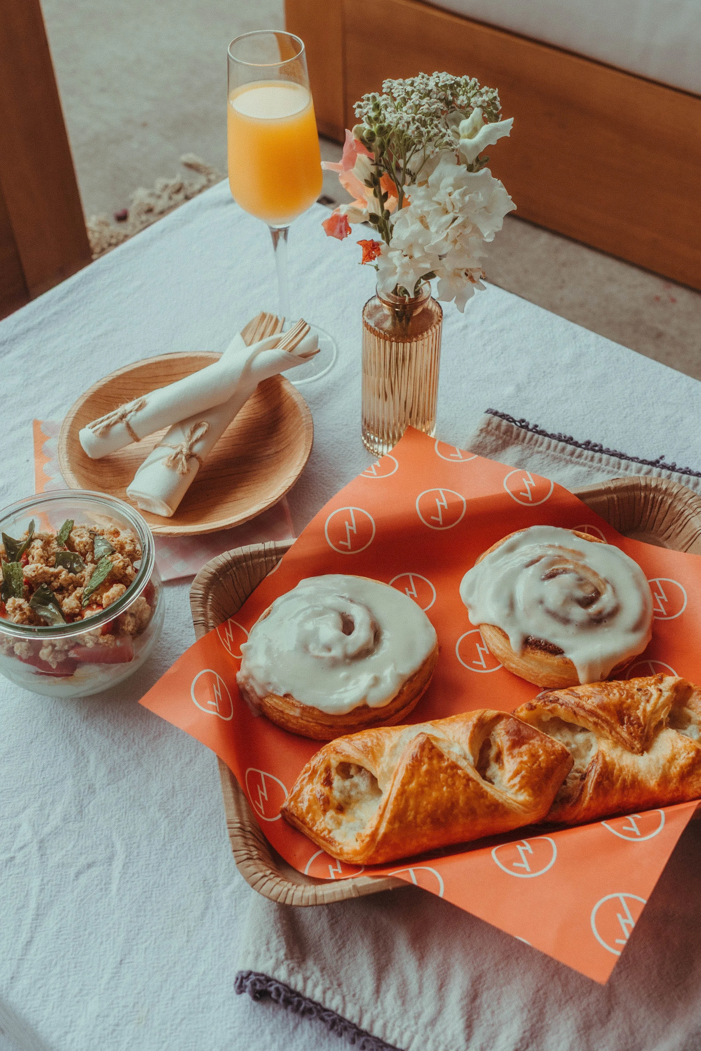 A breakfast table with cinnamon rolls topped with icing on orange branded paper, a pastry, a bowl of granola with nuts and green fruits, a glass of orange juice, and a small vase with white and pastel pink flowers.