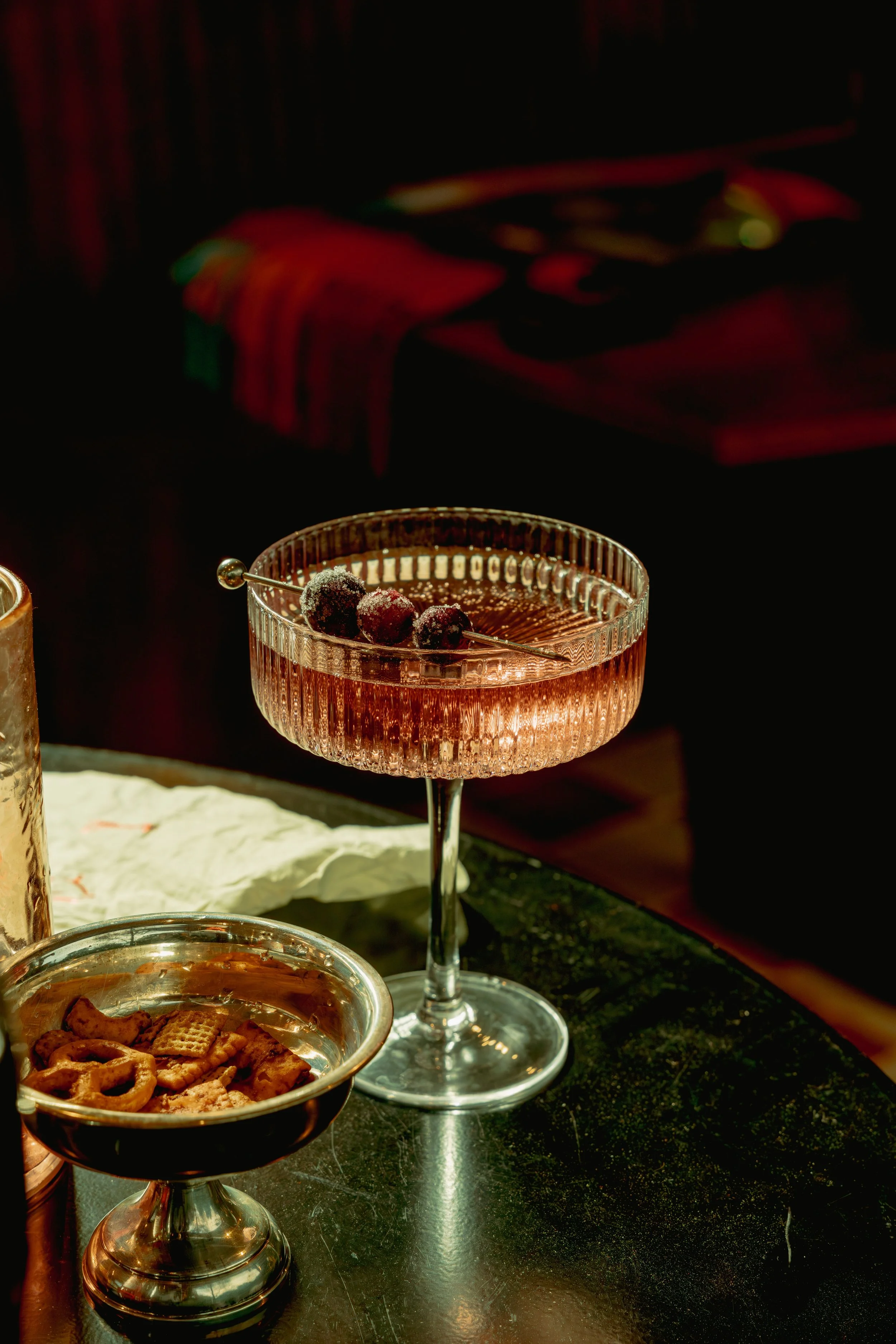 A pink cocktail with blackberries garnished in a vintage glass on a dark bar counter, with a small bowl of crackers nearby.