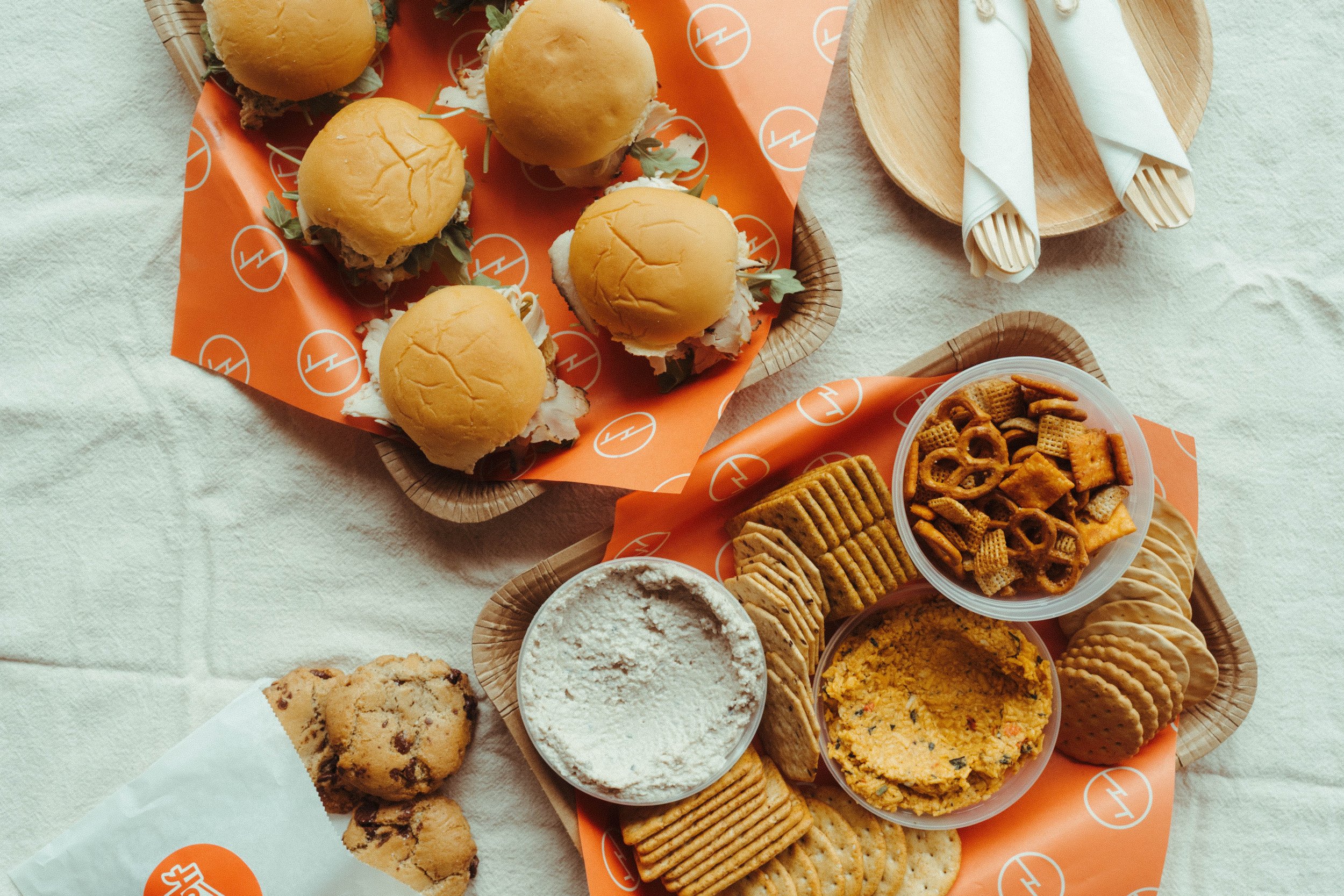 Assorted snacks including mini cheeseburgers, cookies, crackers, chips, and dip on a table with orange branded paper