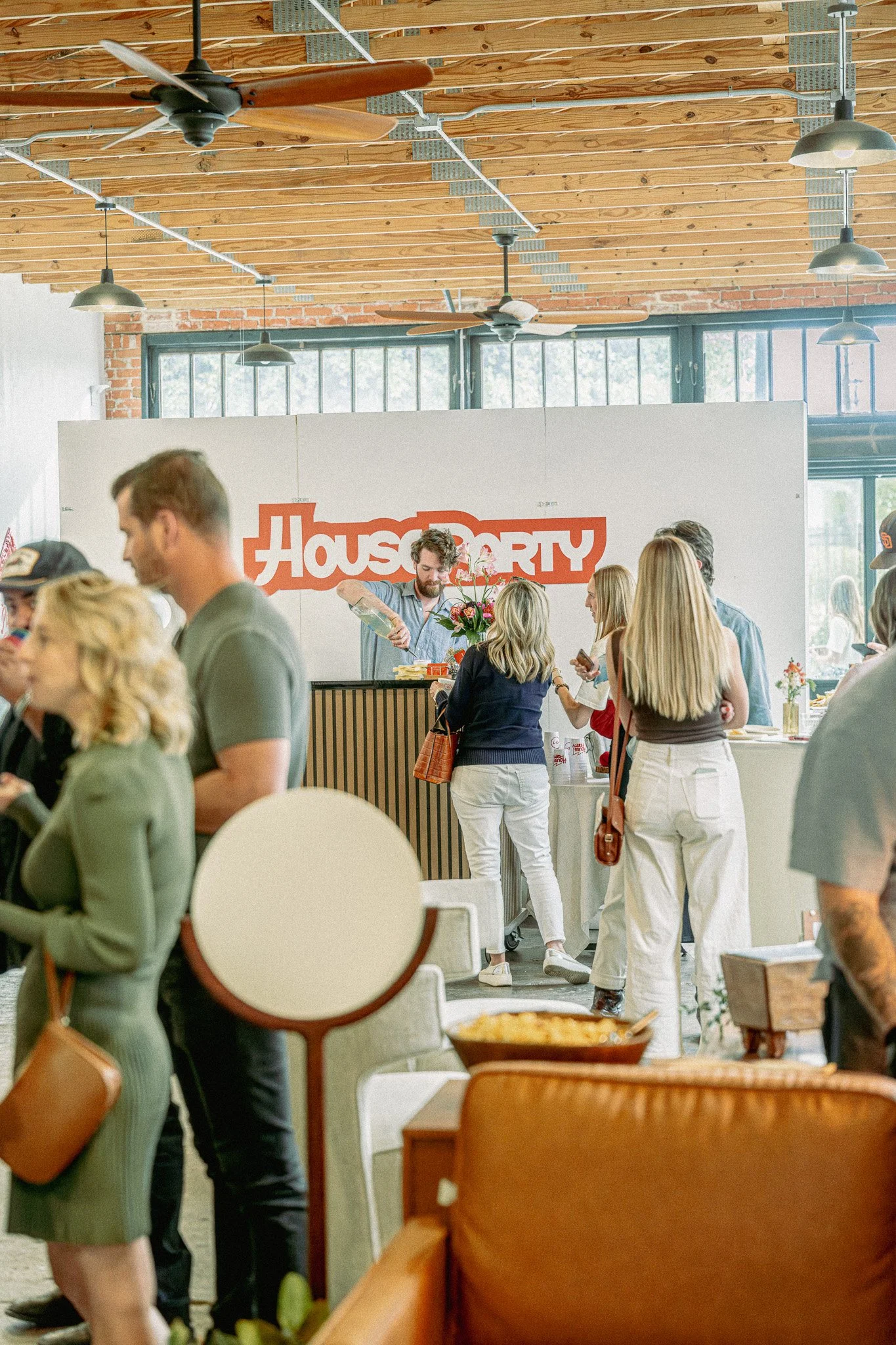 People gathering inside a restaurant with wooden ceiling and large windows. A man is serving food at the counter, and there are several groups of people socializing, some standing and some sitting at tables. The restaurant has a casual, modern decor 