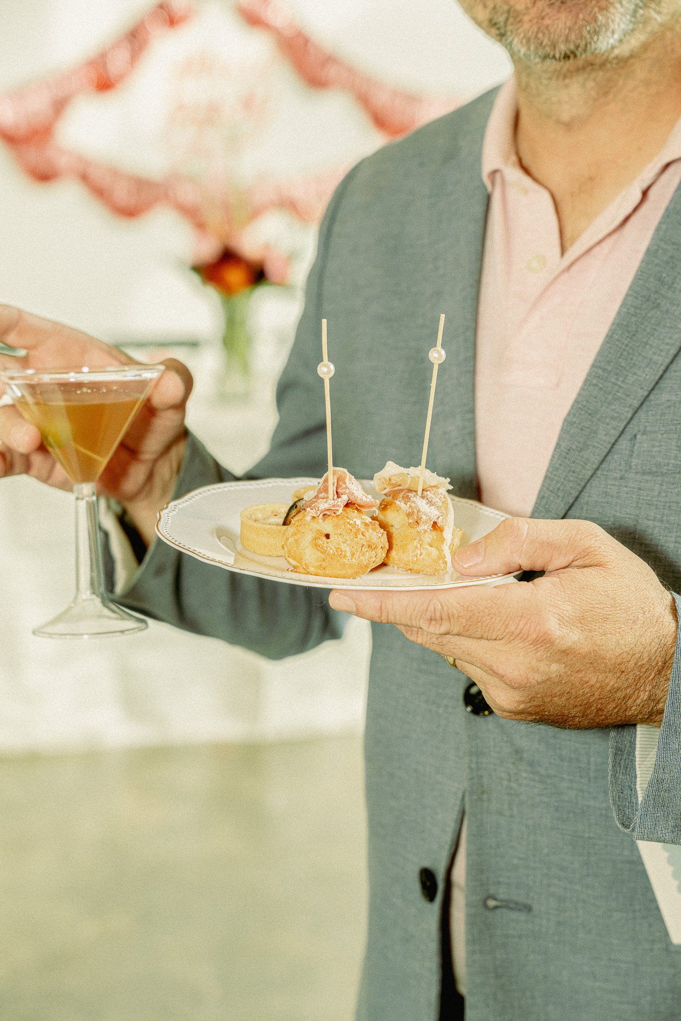 A man in a gray blazer and pink shirt holding a plate with appetizers. Another person in the background is pouring a drink into a martini glass, with a blurred pink flower bouquet in the background.