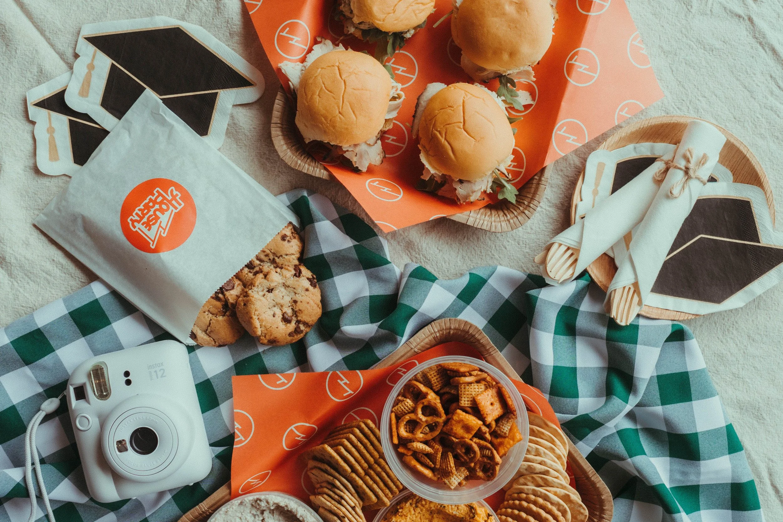A picnic spread with mini sandwiches, chocolate chip cookies, and various snacks on a table with a green and white checkered cloth, orange paper with white circular logos, and a white instant camera.