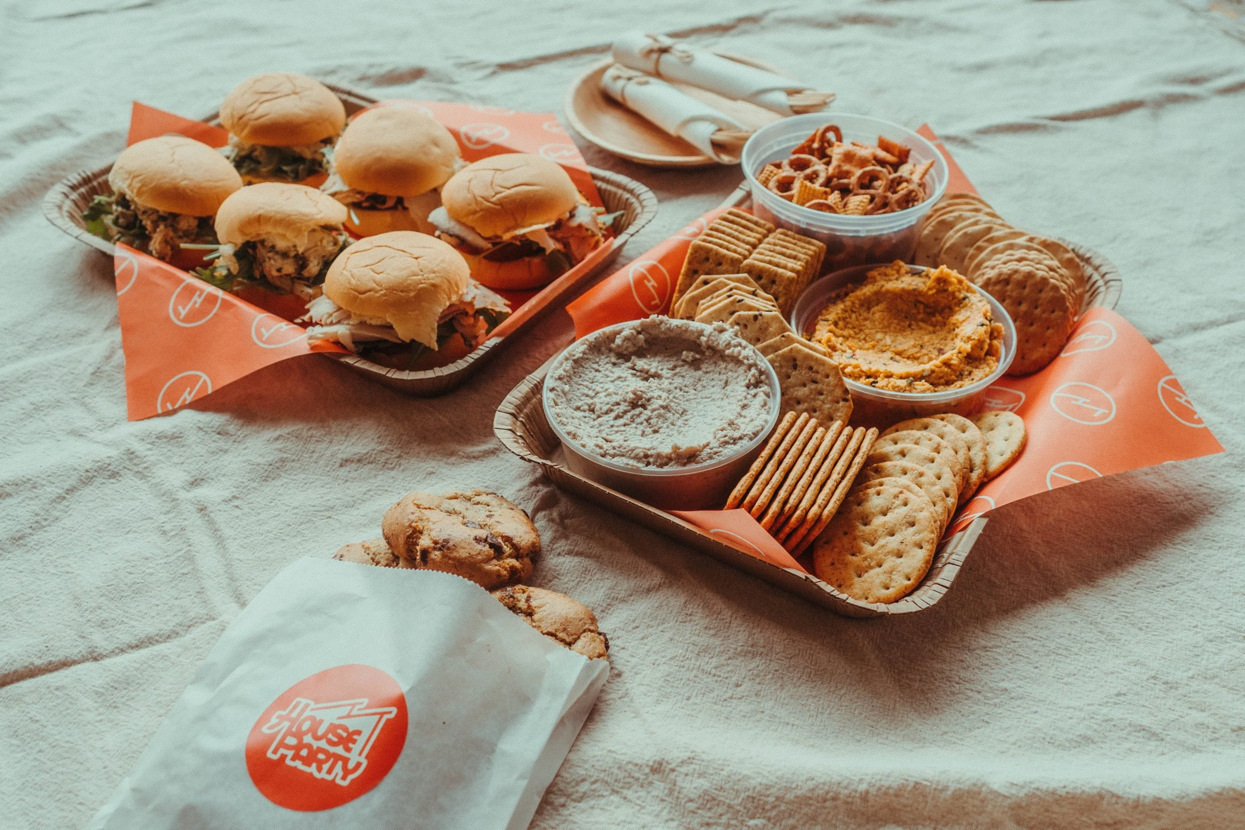A close-up view of a fast food picnic spread on a beige blanket, featuring a tray with pulled pork sandwiches, a cup of snack mix, a bowl of hummus or dip with crackers, and a paper bag of fries with a logo, all set outdoors.