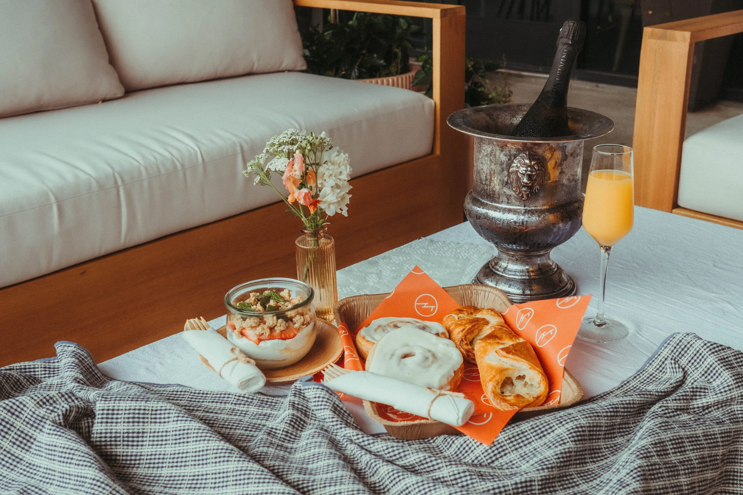 A cozy indoor setting with a table full of breakfast pastries, a bowl of yogurt with fruit, a glass of orange juice, and a small vase with flowers. In the background, there is a white couch, a plant, and a bucket with a bottle of champagne or sparkling wine.