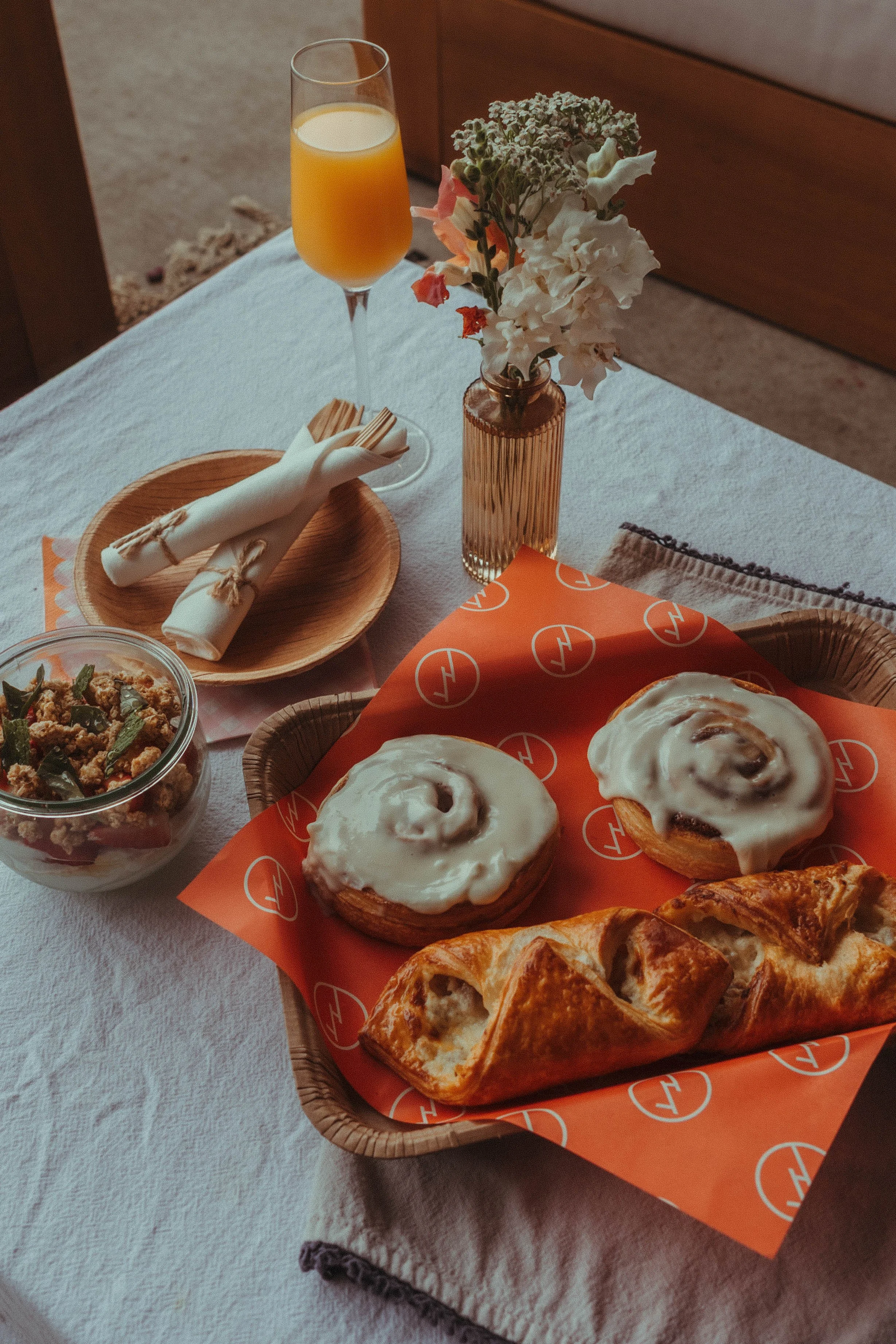 A wooden table set with a glass of orange juice, a vase of white and pink flowers, a bowl of salad, three cinnamon rolls with white icing on a red liner, and slices of cinnamon bread.