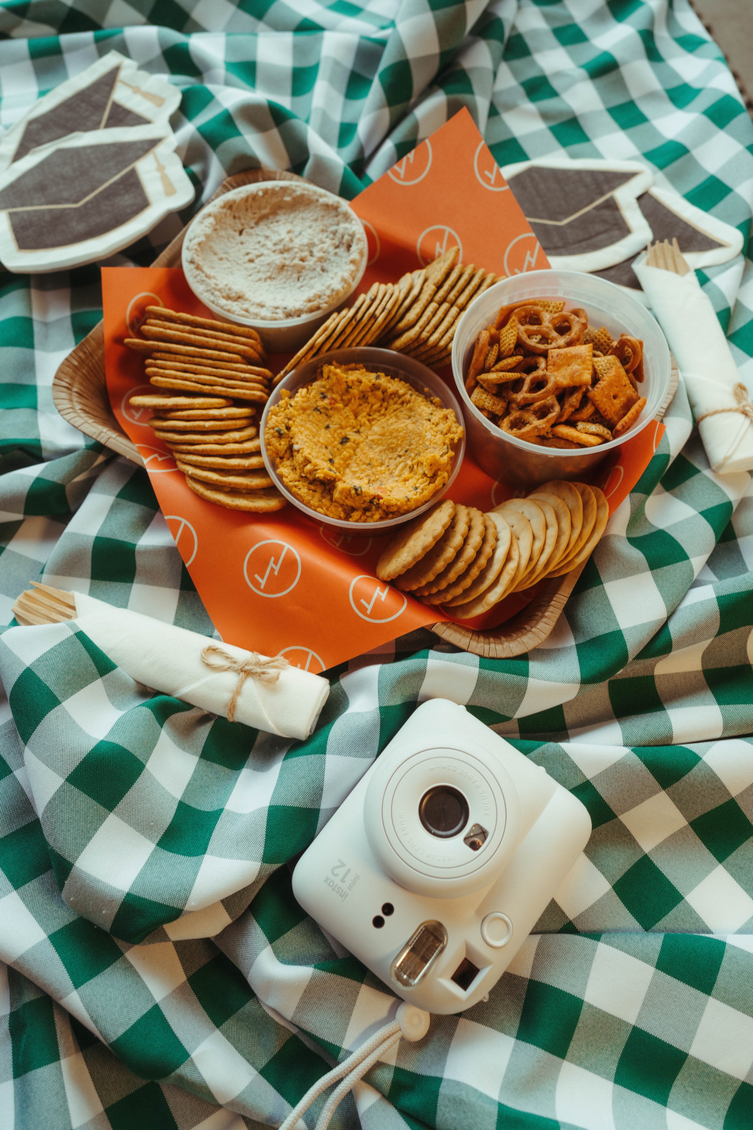 A tray of snack food on a green and white checkered tablecloth. The tray contains a bowl of powdered dip, a bowl of seasoned Chex mix, a bowl of cheese spread, and a variety of crackers. A disposable camera is on a napkin next to the tray.