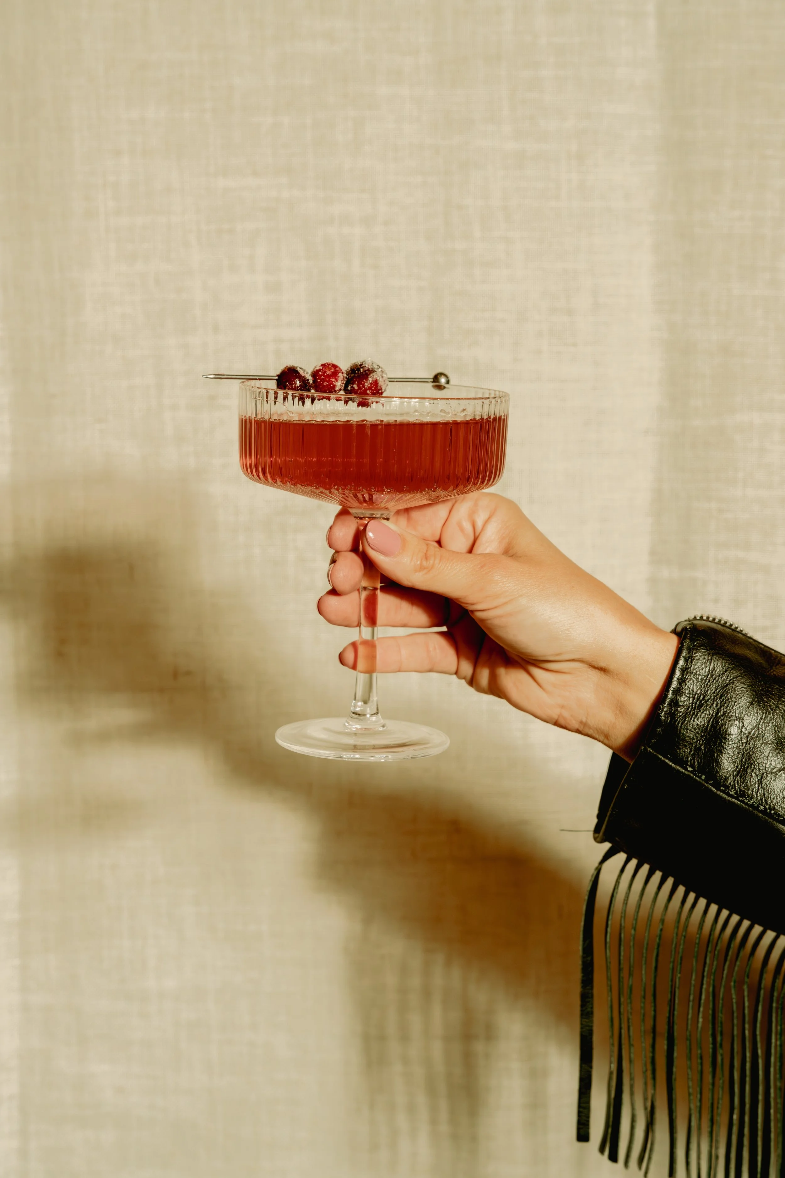 A hand holding a glass of red cocktail garnished with berries and a skewer, against a plain background.
