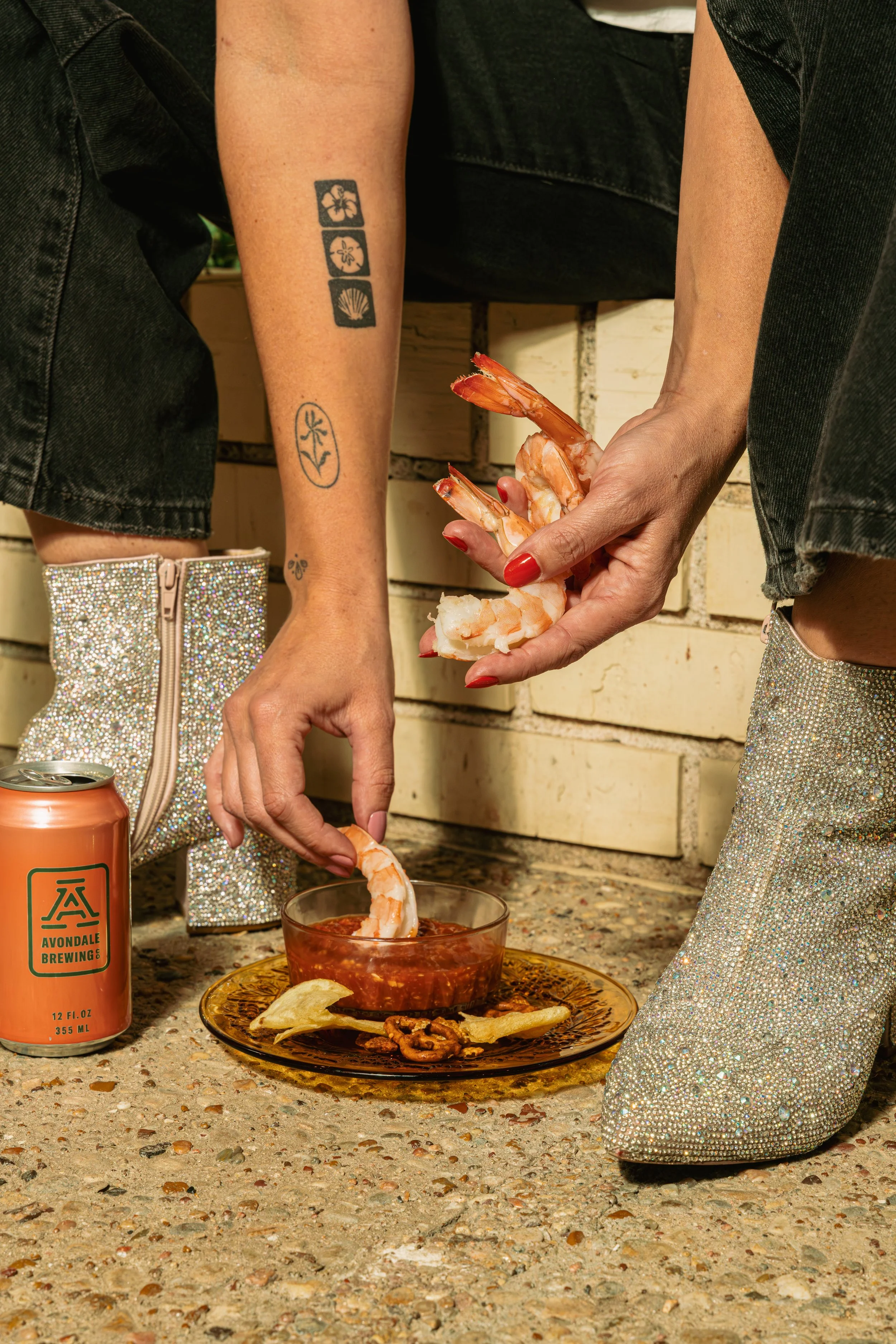 Person sitting on a ledge with glittery boots, dipping cooked shrimp into cocktail sauce on a glass plate with chips and nuts, against a brick wall, with tattoos on arm and a can of beer nearby.