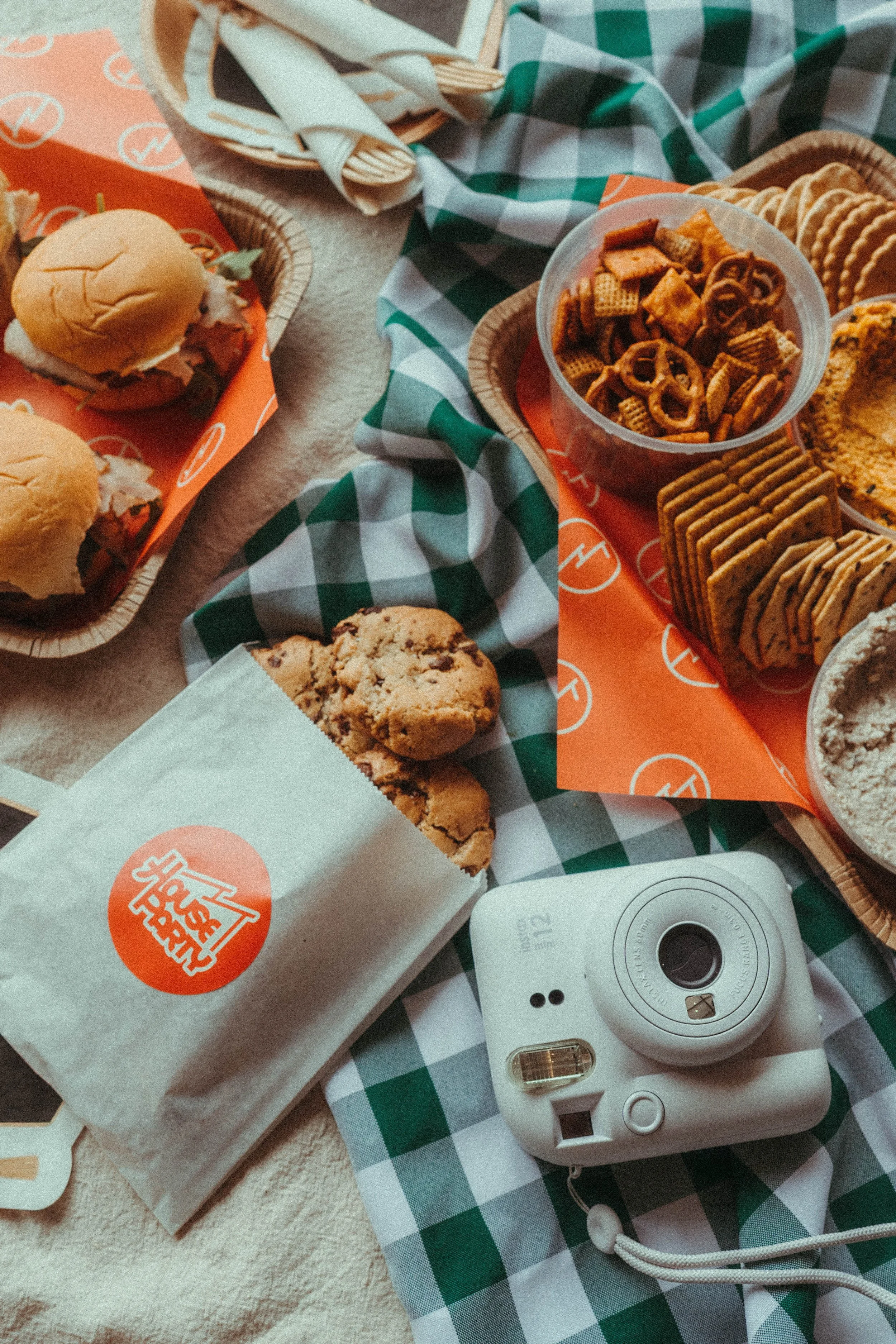 Fast food items including sandwiches, chips, crackers, cookies, and a white instant camera on a checkered tablecloth.