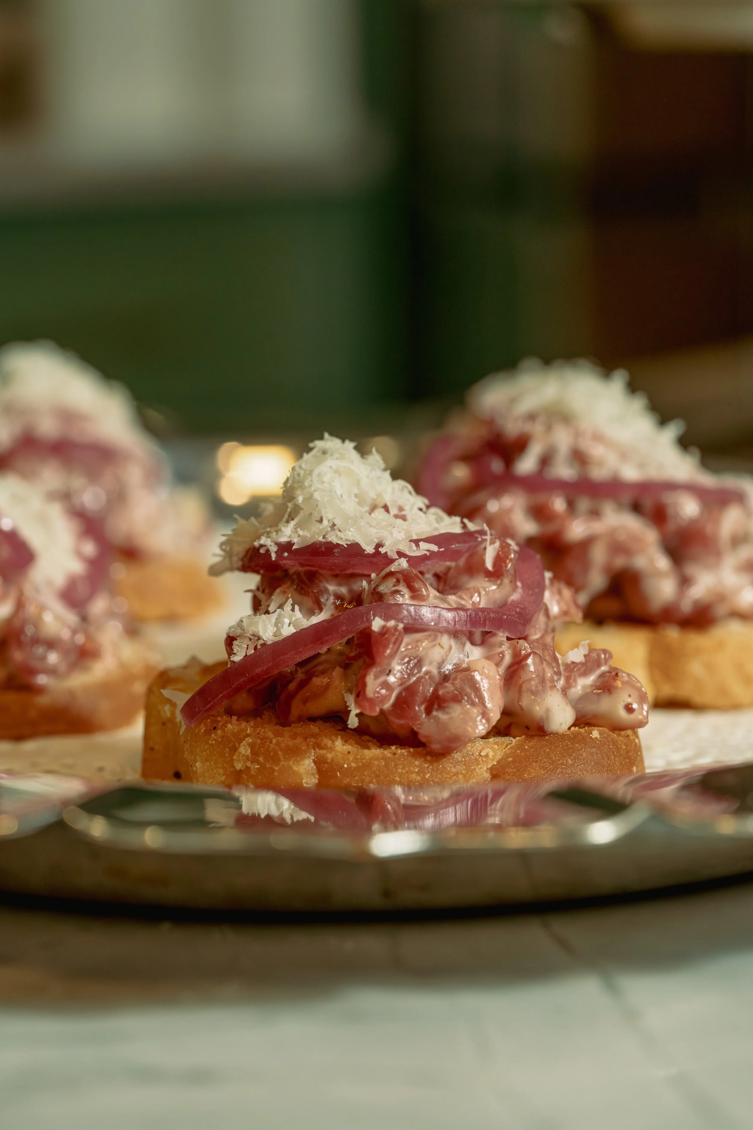 Close-up of an open-faced appetizer with a crispy bread base topped with creamy salad, red onion slices, and shredded cheese, placed on a silver tray.