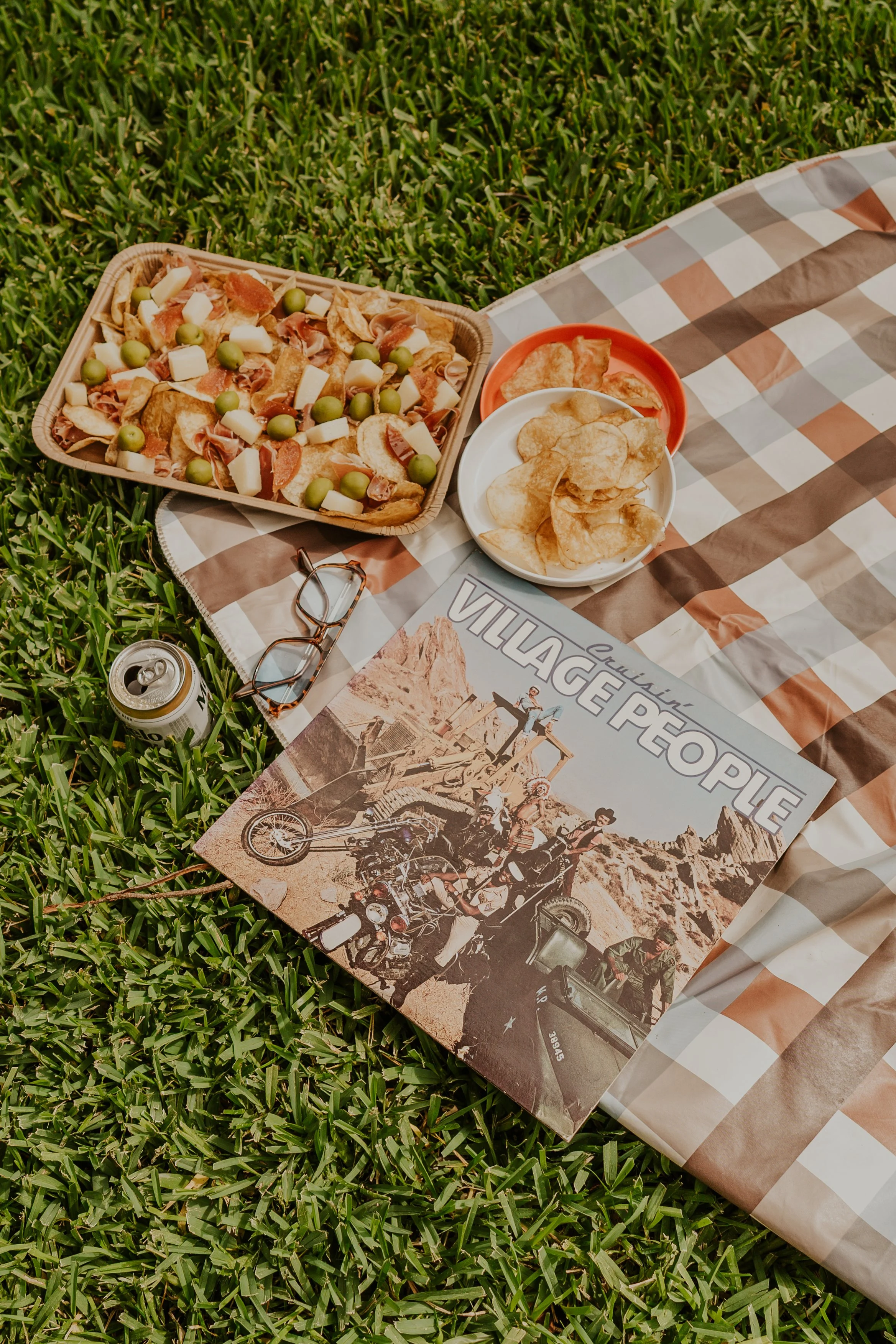 A picnic setup on green grass includes a basket of assorted snacks, two bowls of potato chips, a can of soda, sunglasses, and a magazine with motorcycle riders in a desert landscape.