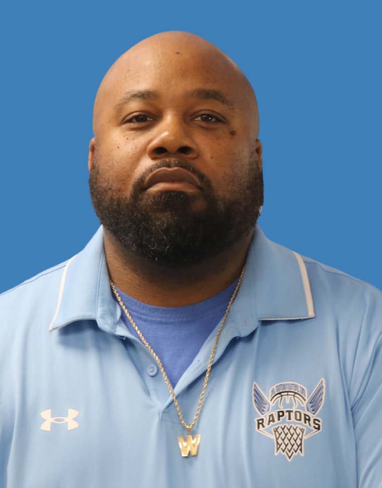 A smiling woman with shoulder-length black hair, wearing a blue top, standing in a lush green greenhouse or garden with various plants in the background.