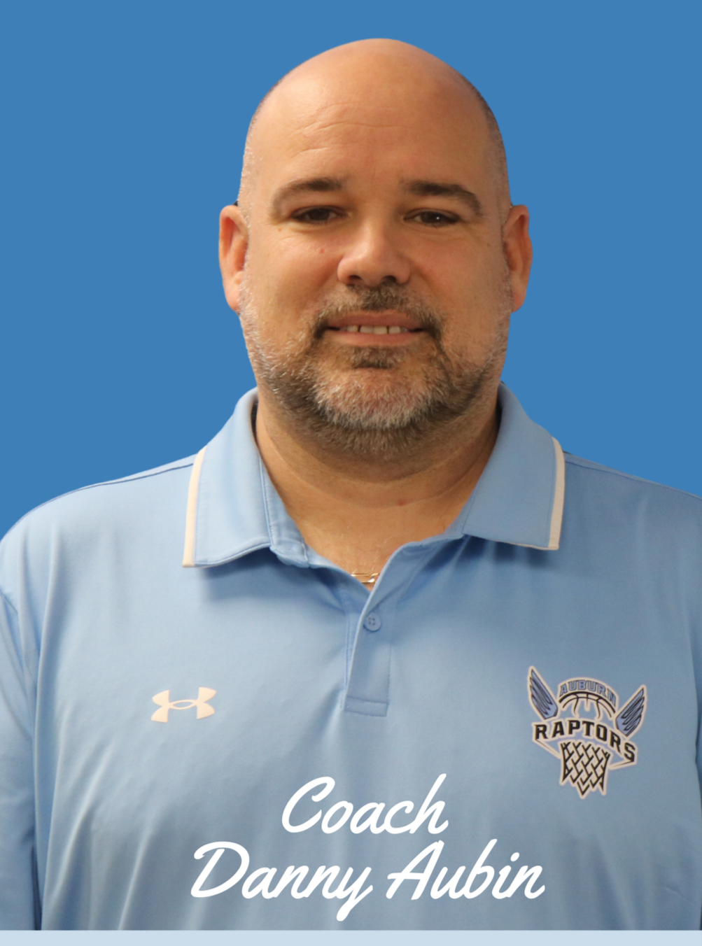 A man with curly dark hair, glasses, and a beard, wearing a black sweater, looking to the side with a slight smile, in a room with a white wall and a plant in the background.