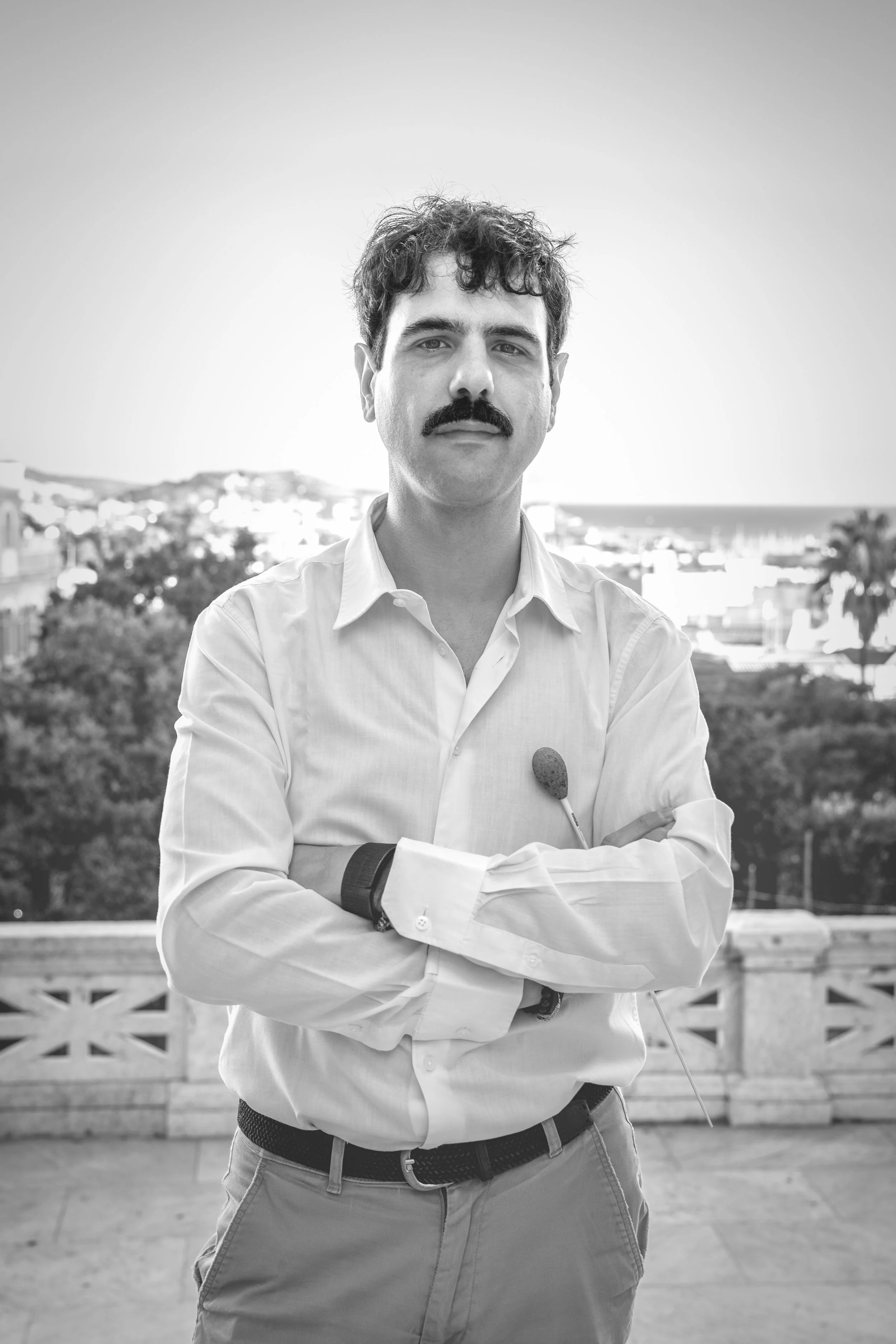 Black and white photo of a man with a mustache and curly hair standing outdoors with arms crossed, wearing a white button-up shirt, a watch, and a belt, with a cityscape, trees, and a body of water in the background.