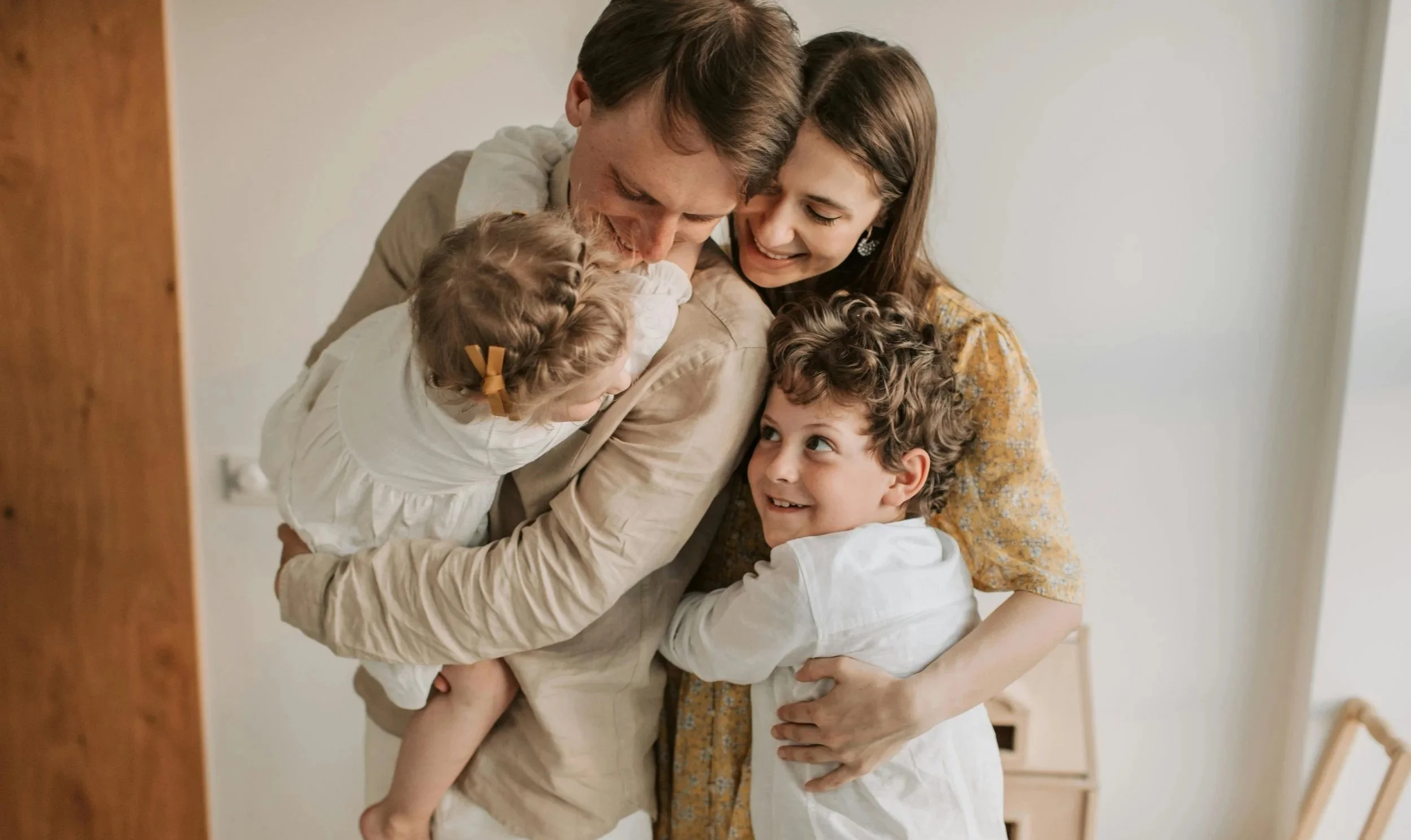 A family of four, a man, woman, and two children, hugging and smiling together in a cozy indoor setting.