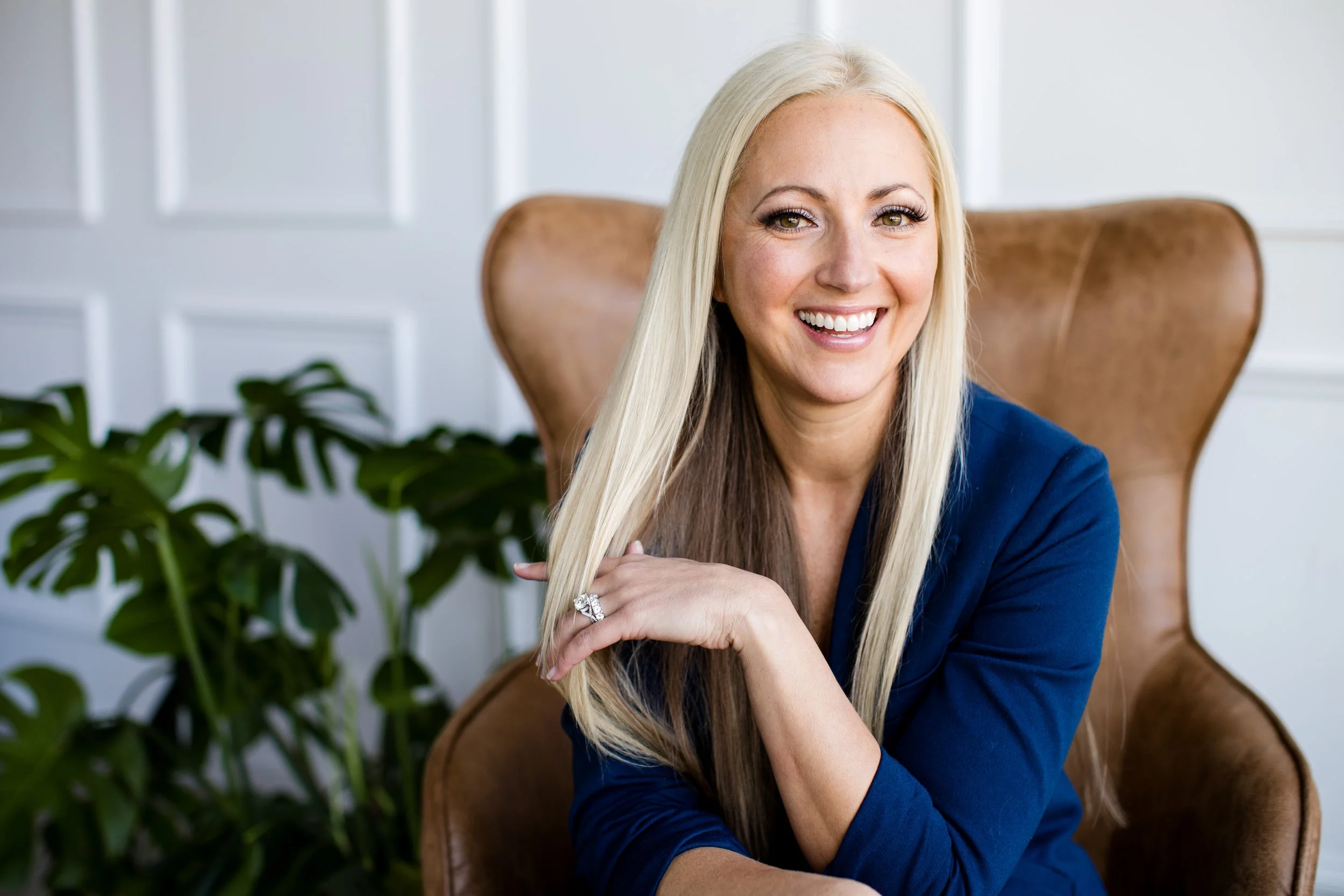 A smiling blonde woman sitting in a brown leather chair, wearing a navy blue top, with a large green plant behind her and a white paneled wall in the background.