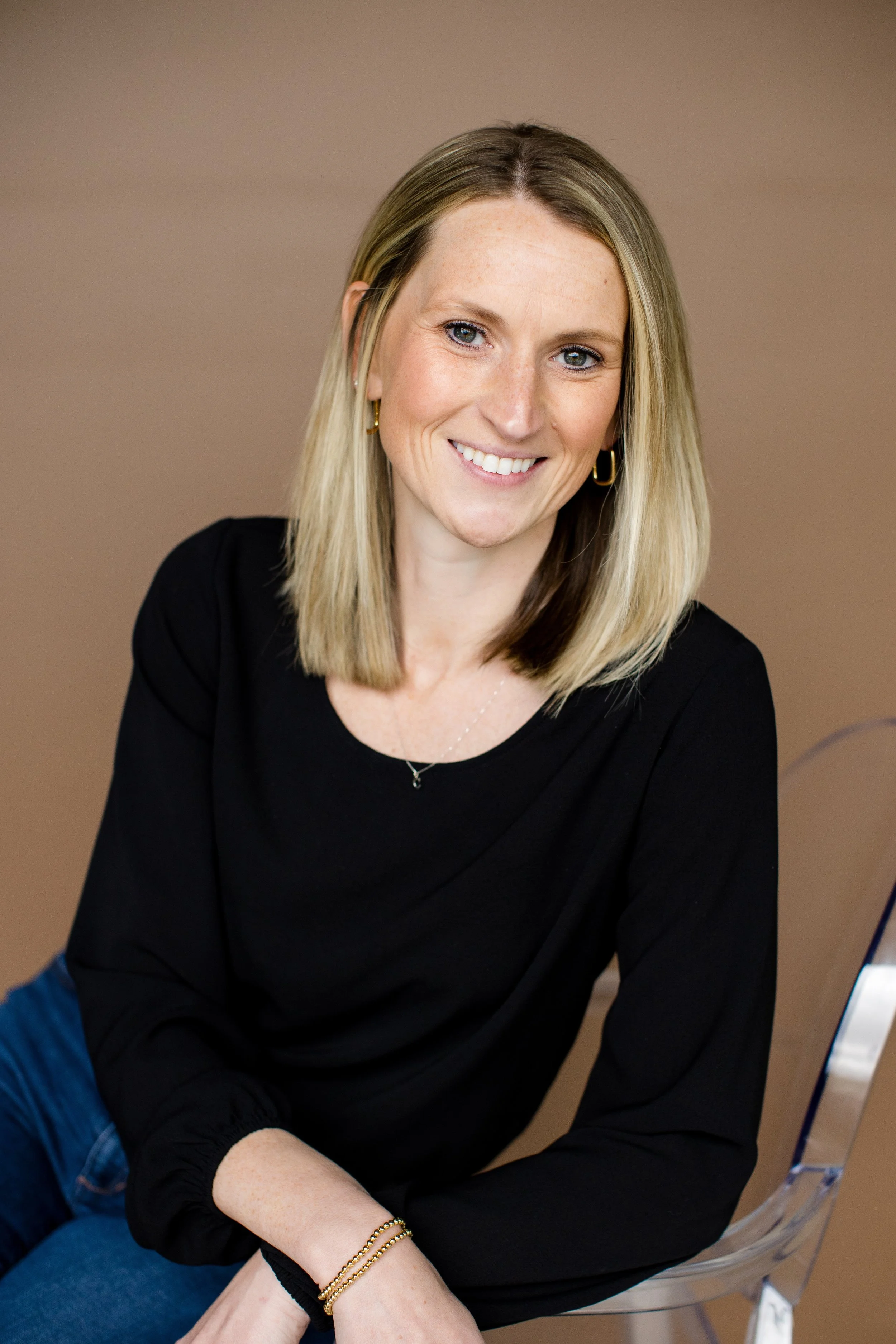 A woman with blonde hair, wearing a black top, sitting on a clear chair, smiling at the camera.