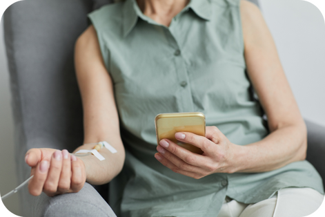 A woman sitting on a couch receiving an intravenous injection in her hand while looking at her phone.