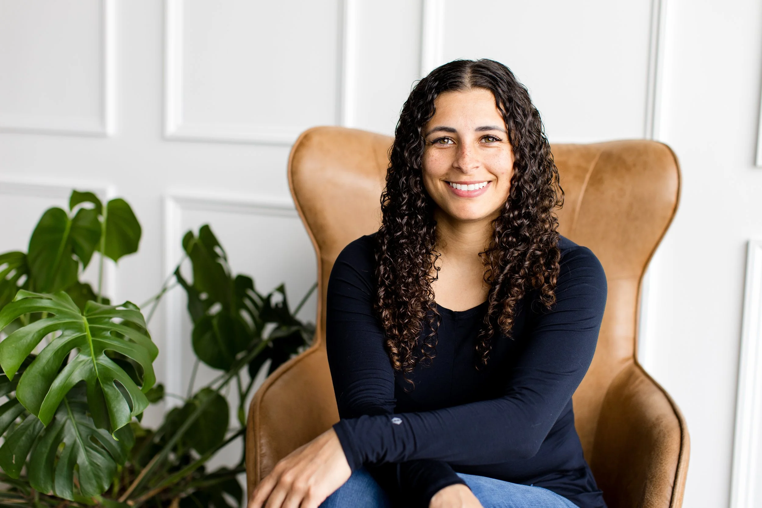 Smiling woman with curly hair sitting in a tan armchair next to green houseplants, in a room with white paneled walls.
