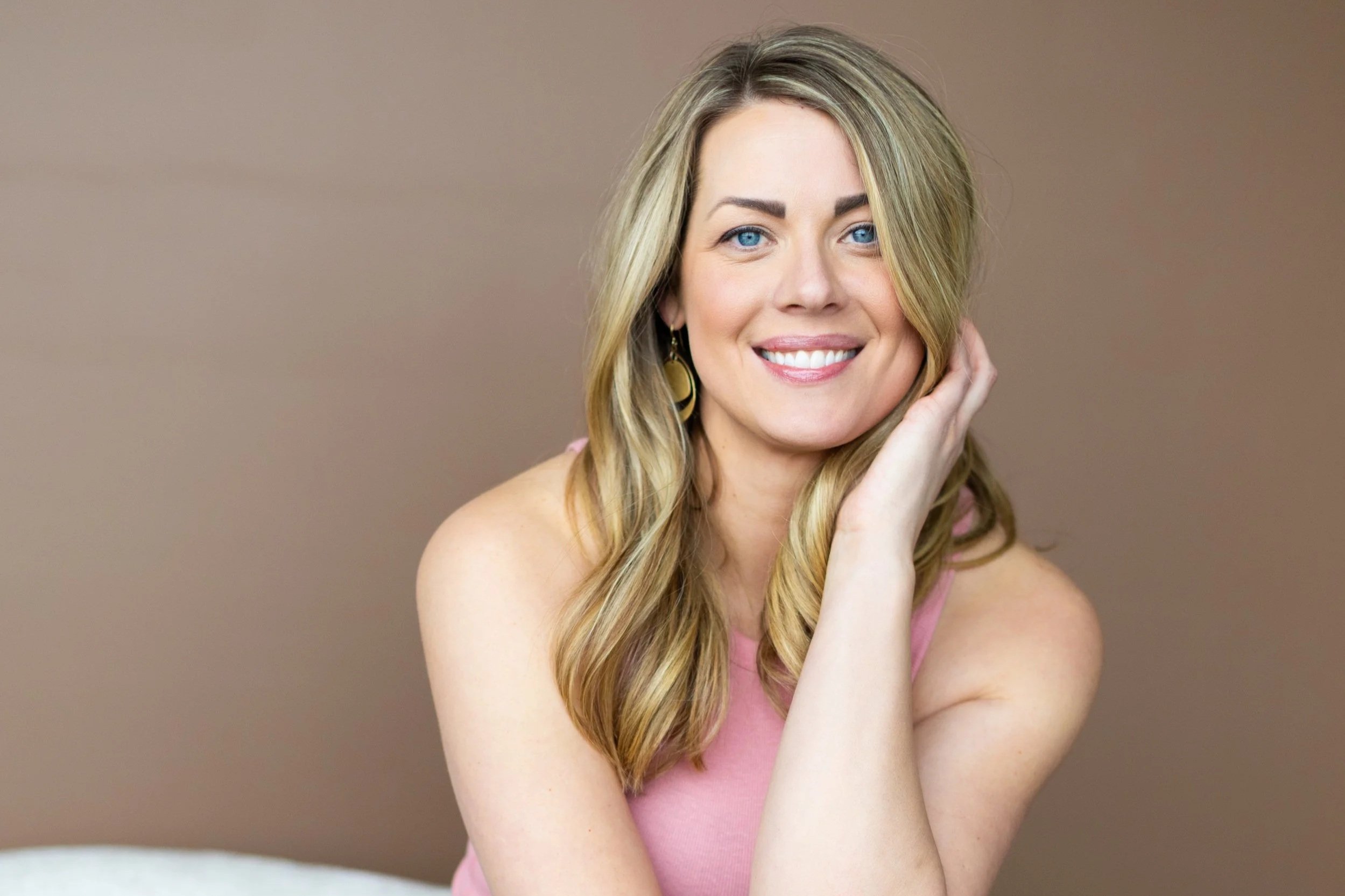 A woman with long blonde hair, wearing a pink sleeveless top and gold hoop earrings, smiling with her right hand touching her face, against a neutral background.