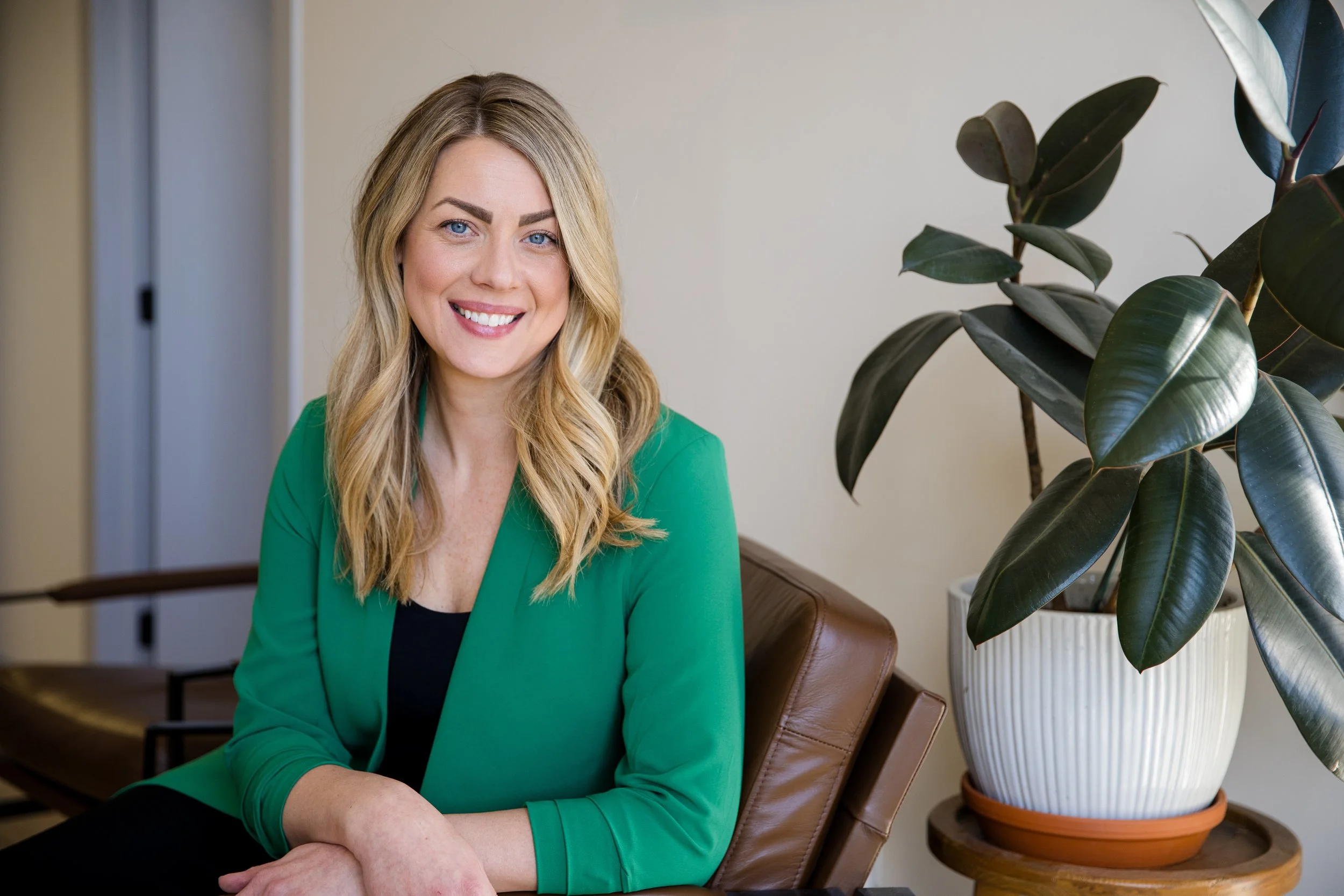 A woman with blonde hair, wearing a green blazer, sitting on a brown leather chair indoors, next to a large potted plant with dark green leaves.