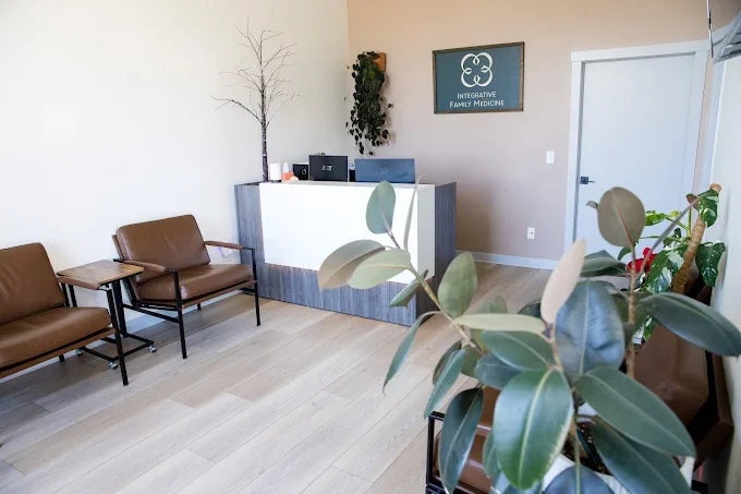 Waiting area with brown chairs and small tables near a reception desk, plants, and a logo that reads 'Integrative Family Medicine' on the wall.