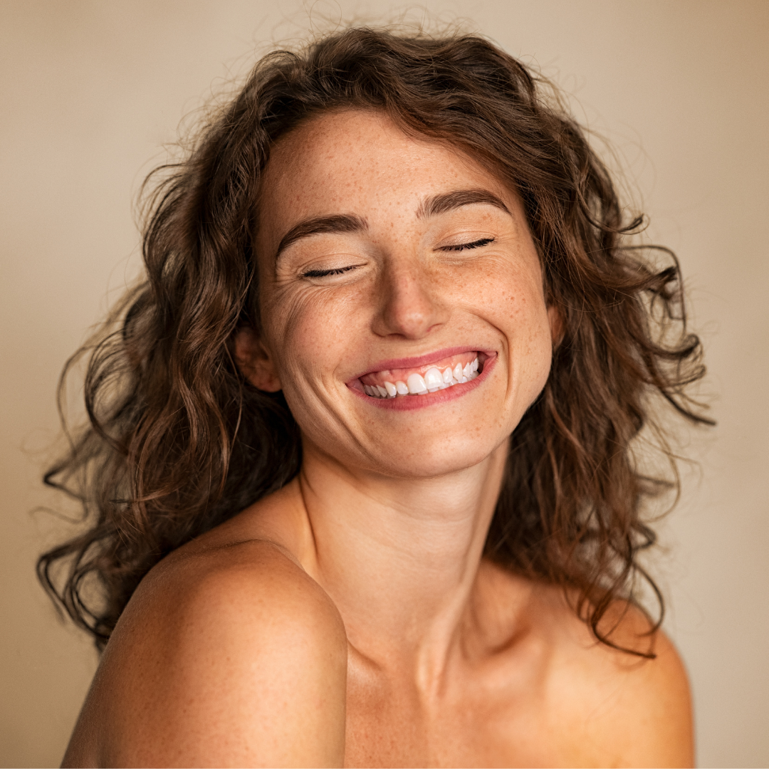 A woman with curly brown hair and freckles, smiling with her eyes closed, showing her teeth.