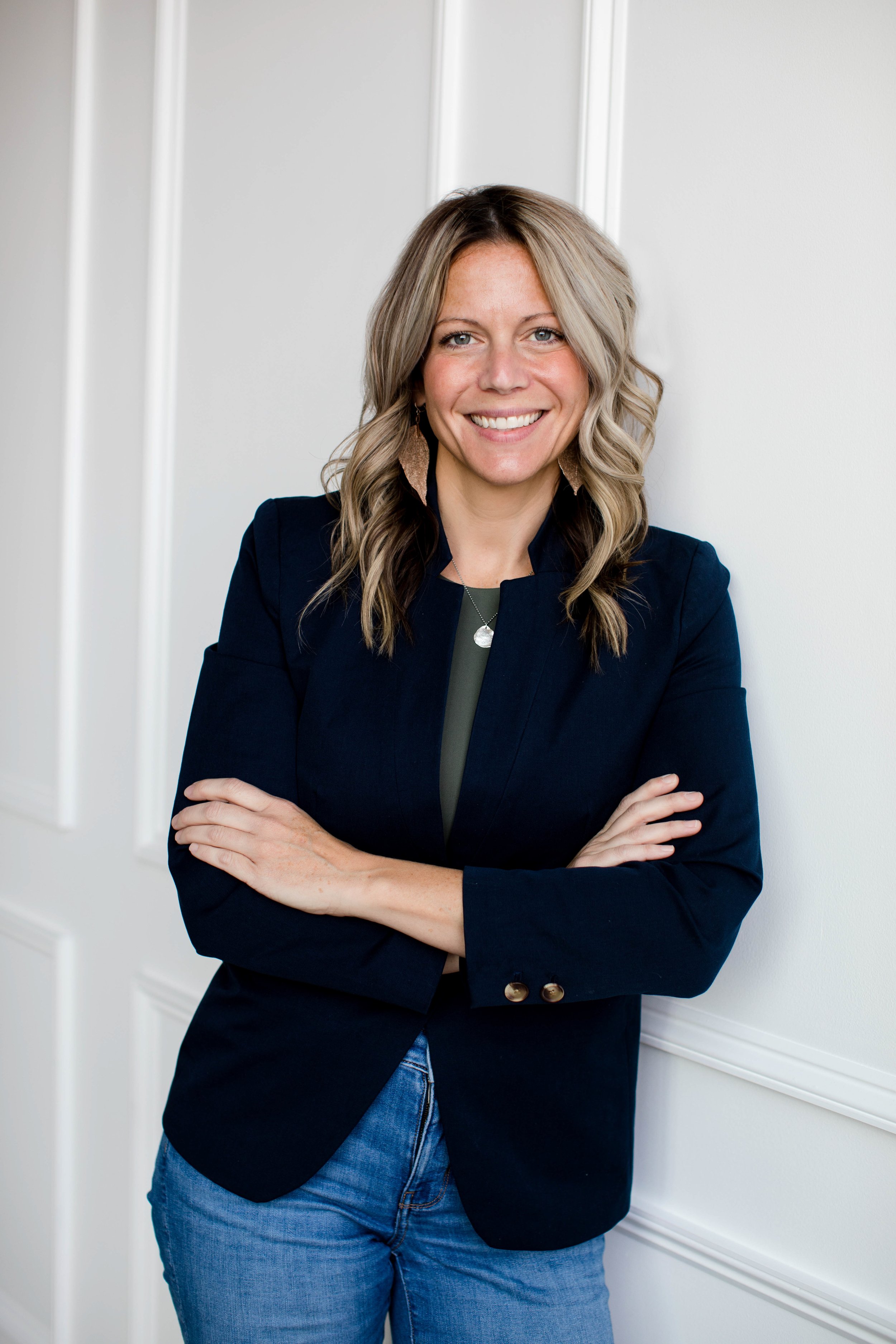 A smiling woman with blonde hair, wearing a black blazer and blue jeans, standing with arms crossed in front of a white wall.