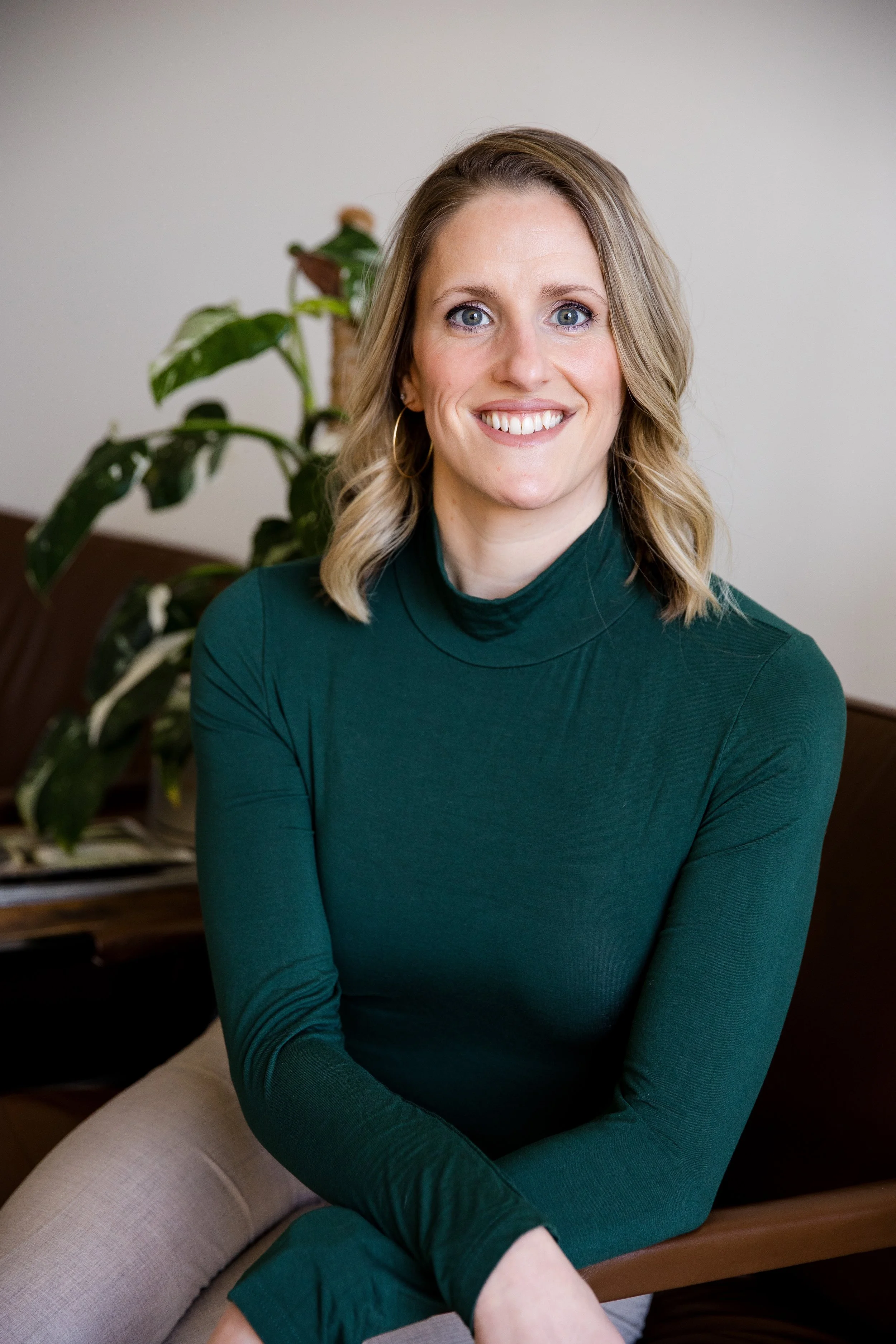 A woman with blonde hair, blue eyes, wearing a green turtleneck, sitting on a bench near a plant, smiling.