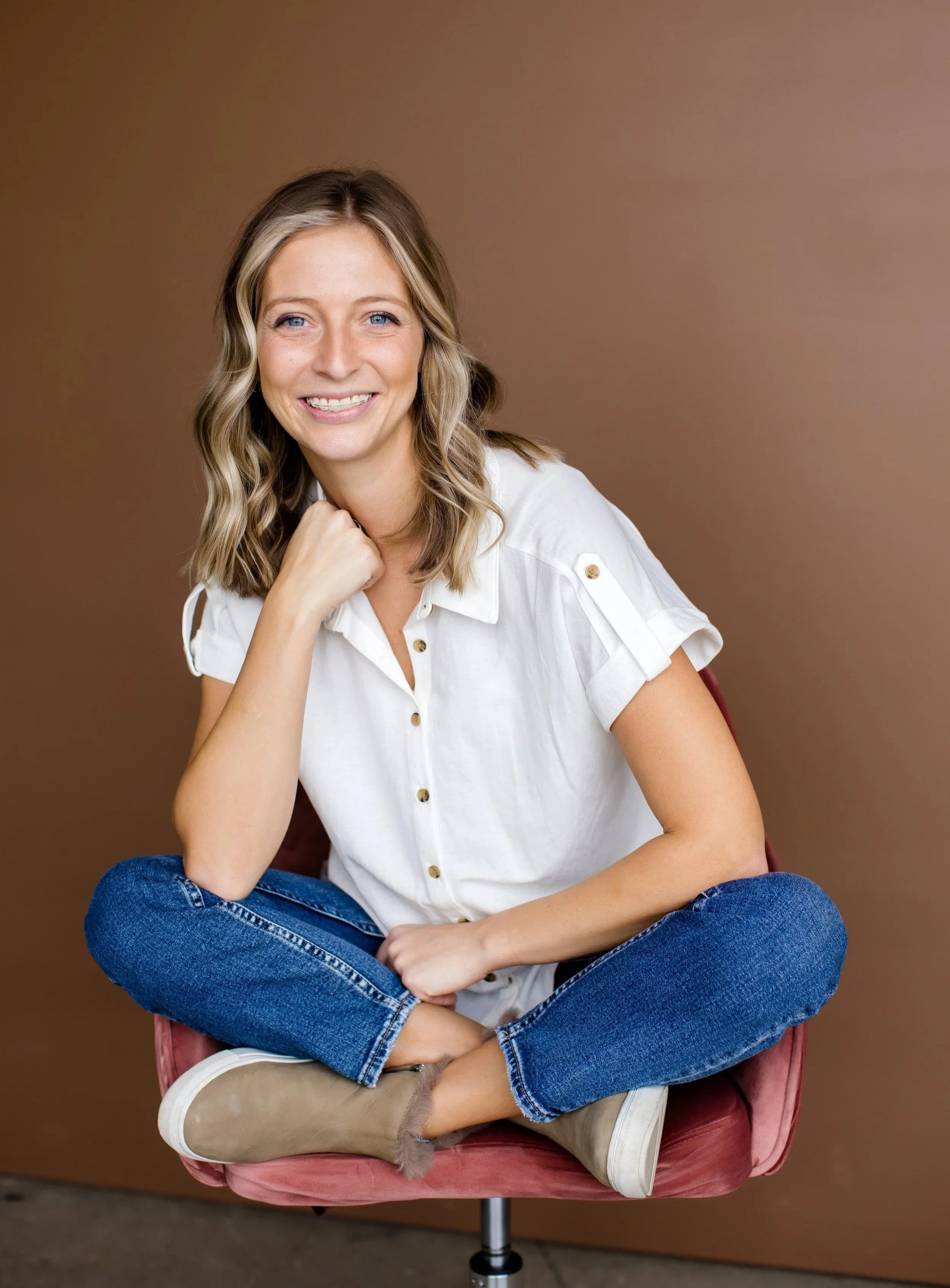 A woman with shoulder-length wavy blonde hair smiling, seated cross-legged on a pink chair against a brown background, wearing a white short-sleeved button-up shirt, blue jeans, and beige ankle boots.