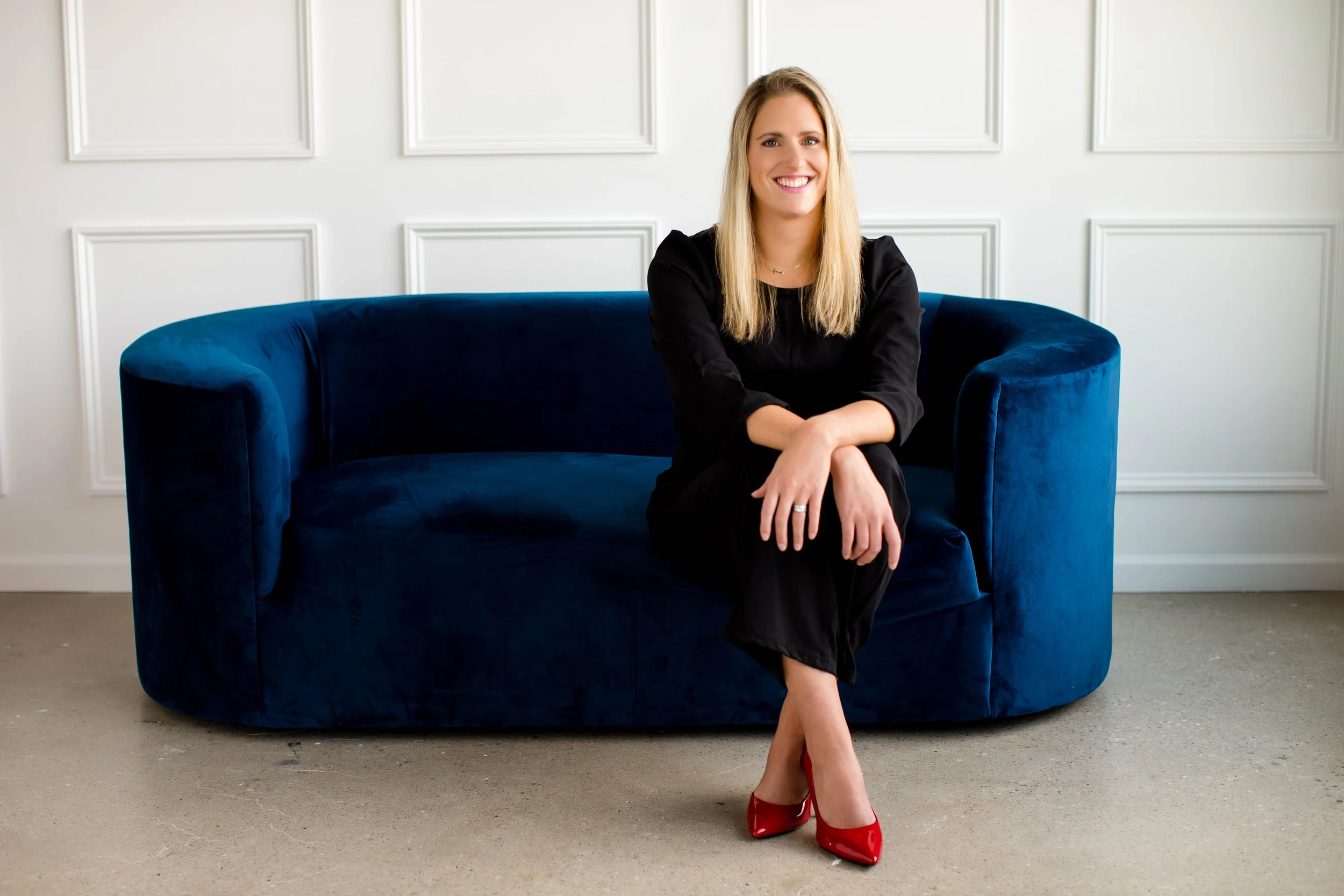 A smiling woman with blonde hair in a black outfit and red high heels sitting on a blue velvet sofa in a white room.