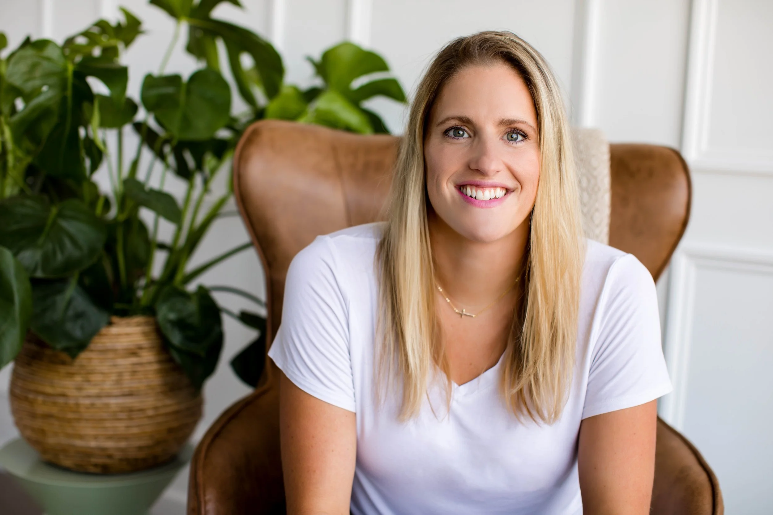 A smiling woman with blonde hair sitting on a brown leather armchair in front of a large green potted plant and a white wall with framed artwork.